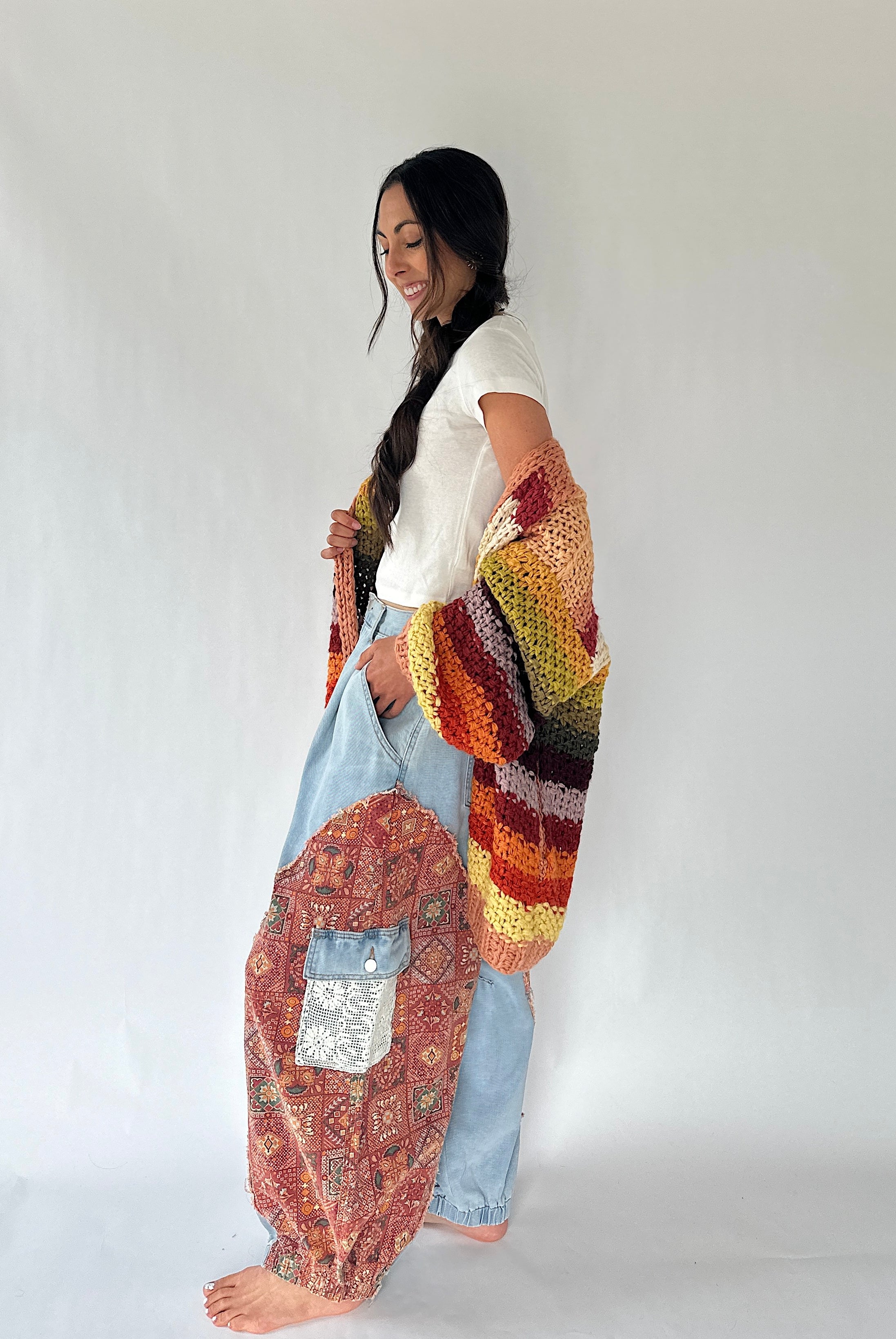 A woman stands barefoot against a plain light gray studio background, wearing a white short-sleeve top, light blue harem-style pants, and a large multicolored crocheted cardigan draped over her arm and around her body. The cardigan has a striped pattern in orange, yellow, green, brown, black, and cream.