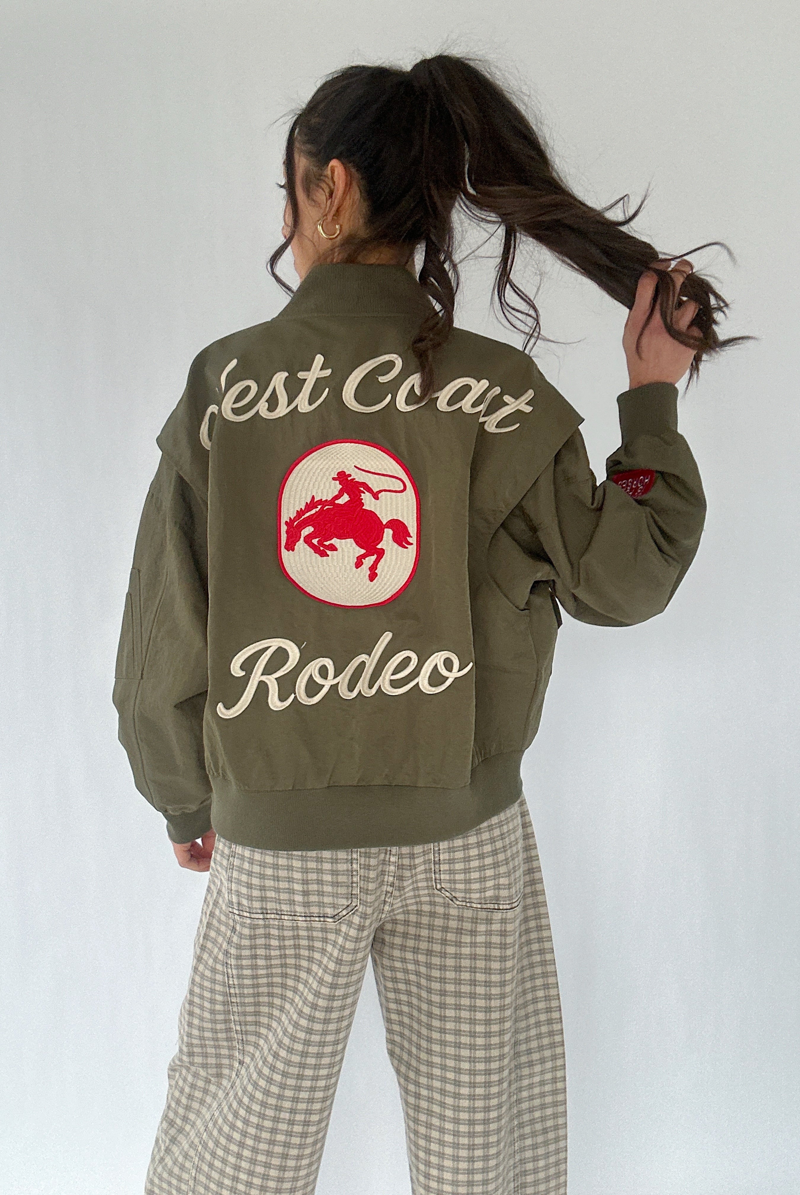 Person wearing a green jacket with 'West Coast Rodeo' text and cowboy logo on the back, standing against a white background.