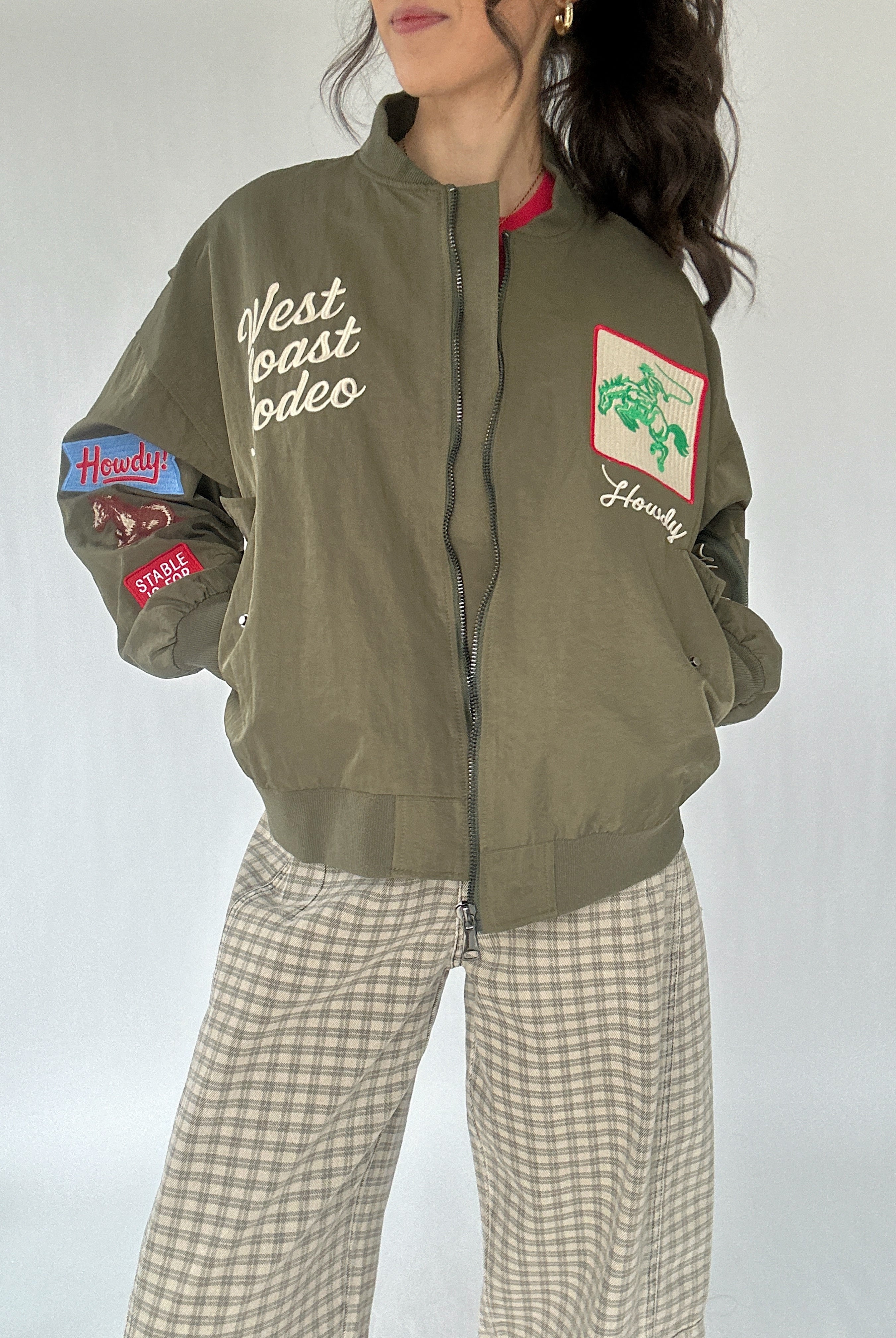 Person wearing a green bomber jacket with western patches and text, standing against a white background