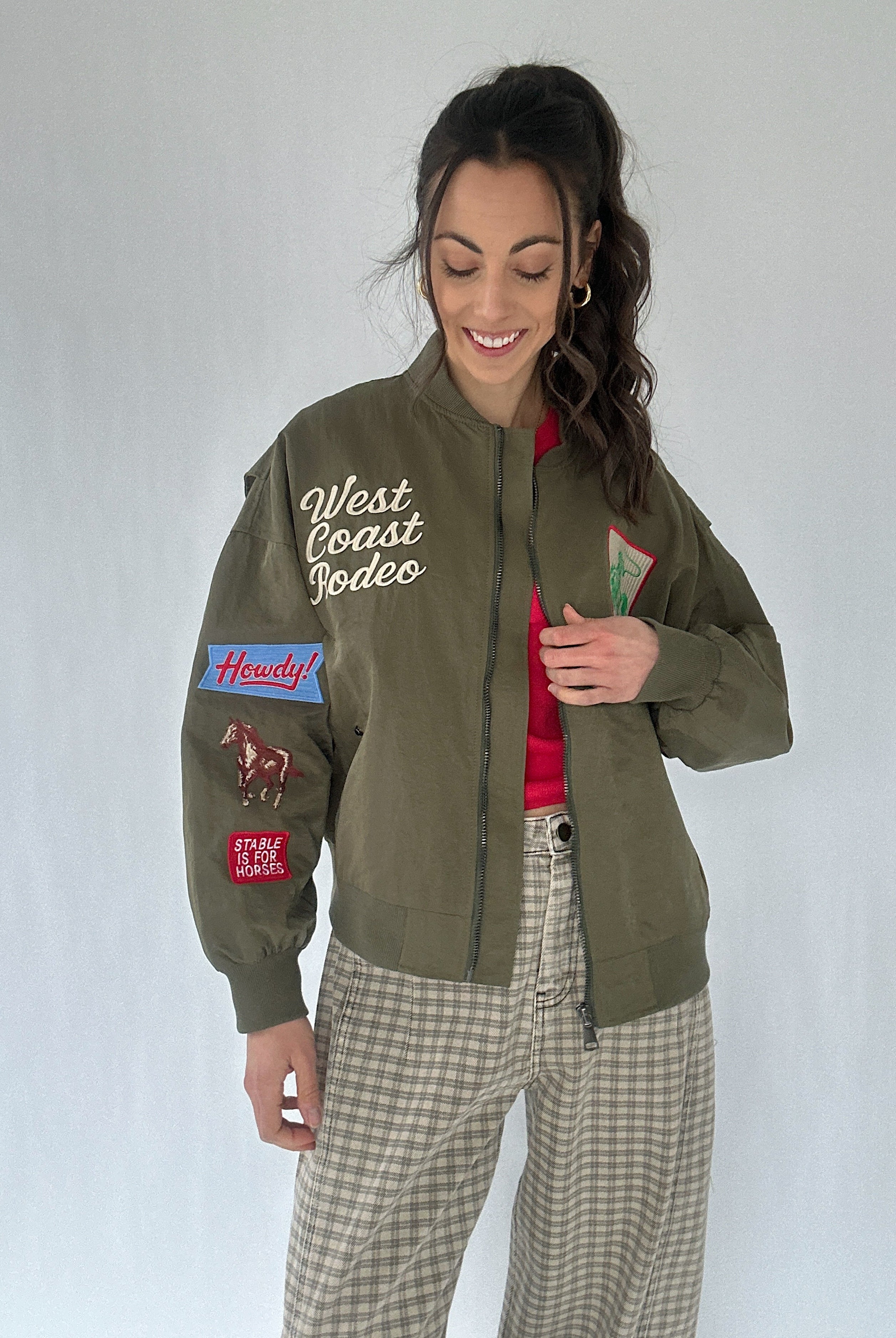 Woman wearing a green jacket with patches and text, standing against a plain background