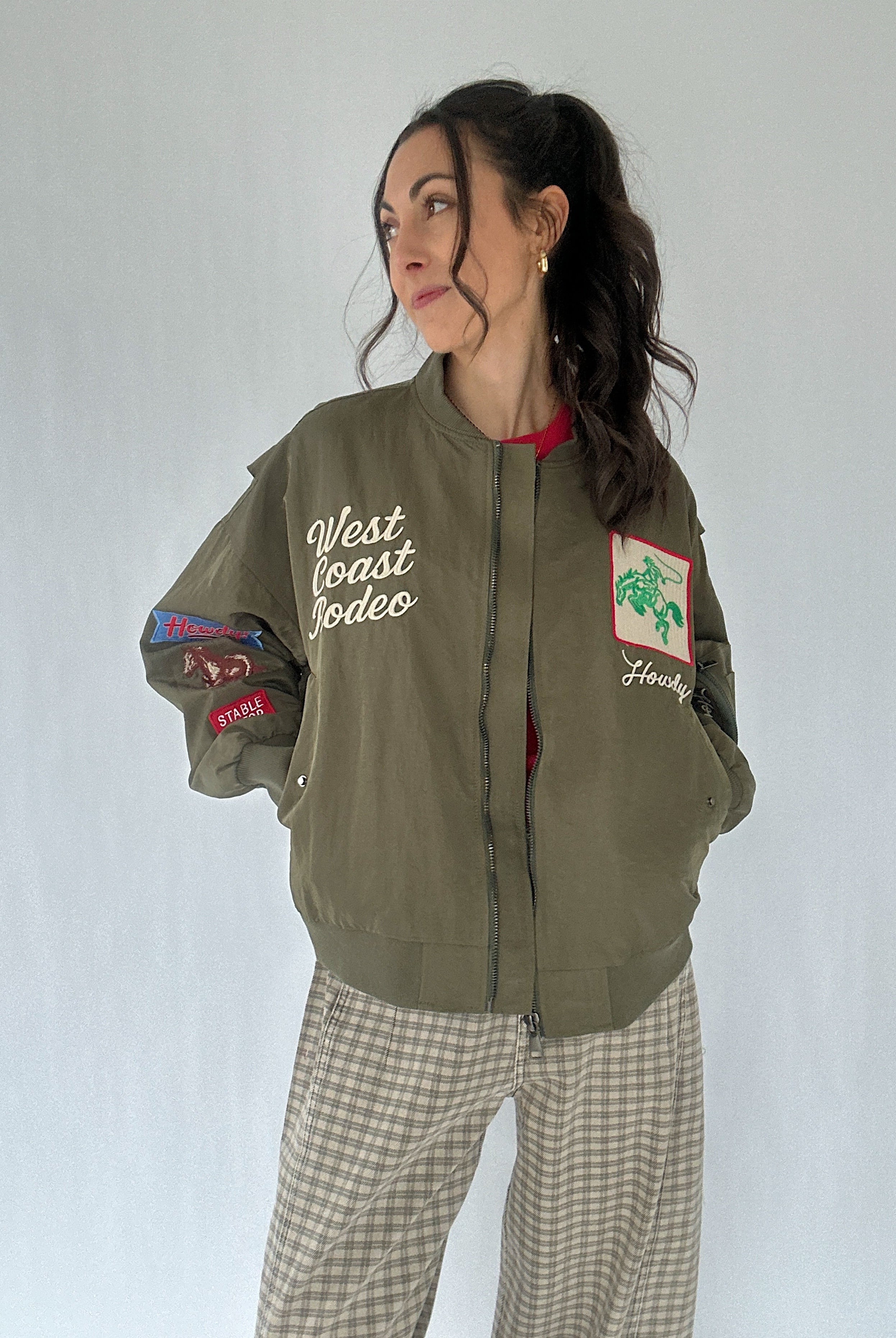 Woman wearing a green bomber jacket with patches and text, standing against a plain background