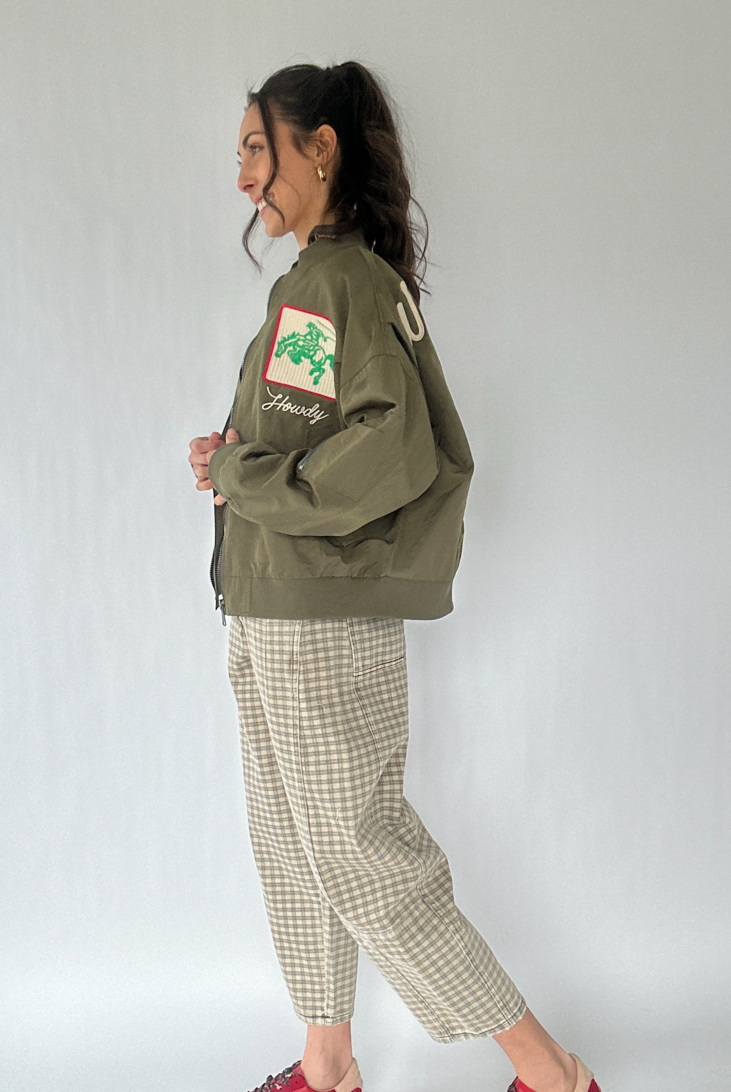 Woman wearing a green jacket with a logo and text, standing against a plain background from side