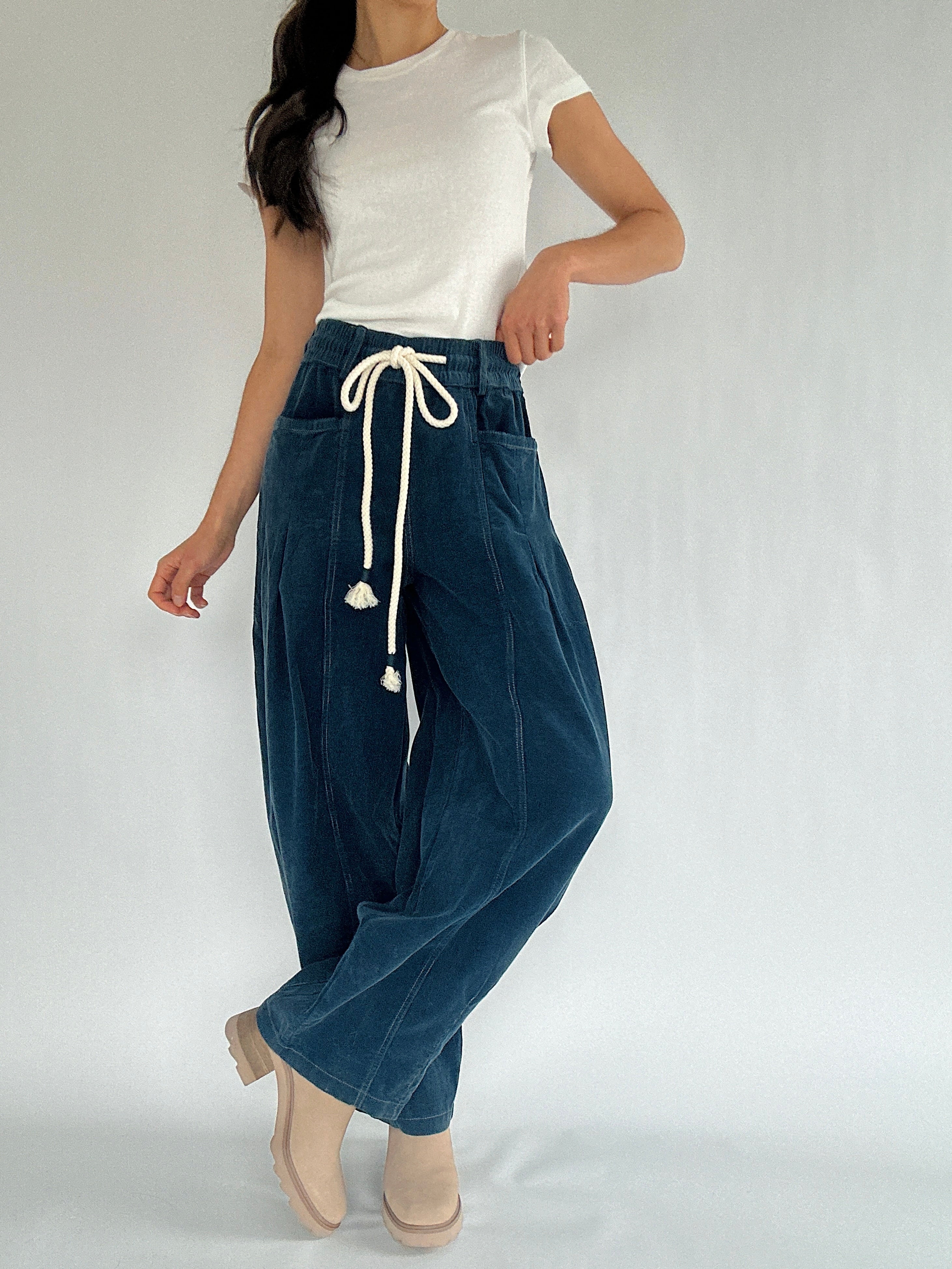 Person wearing blue corduroy barrel drawstring pants with a white top on a plain background with legs crossed