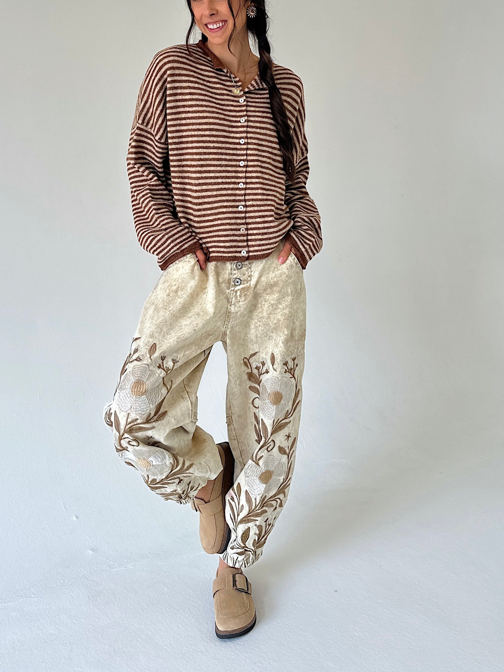 A woman modeling a brown and beige striped long-sleeve button-up shirt with cream-colored harem pants featuring floral embroidery, paired with tan suede boots, against a plain light gray background.