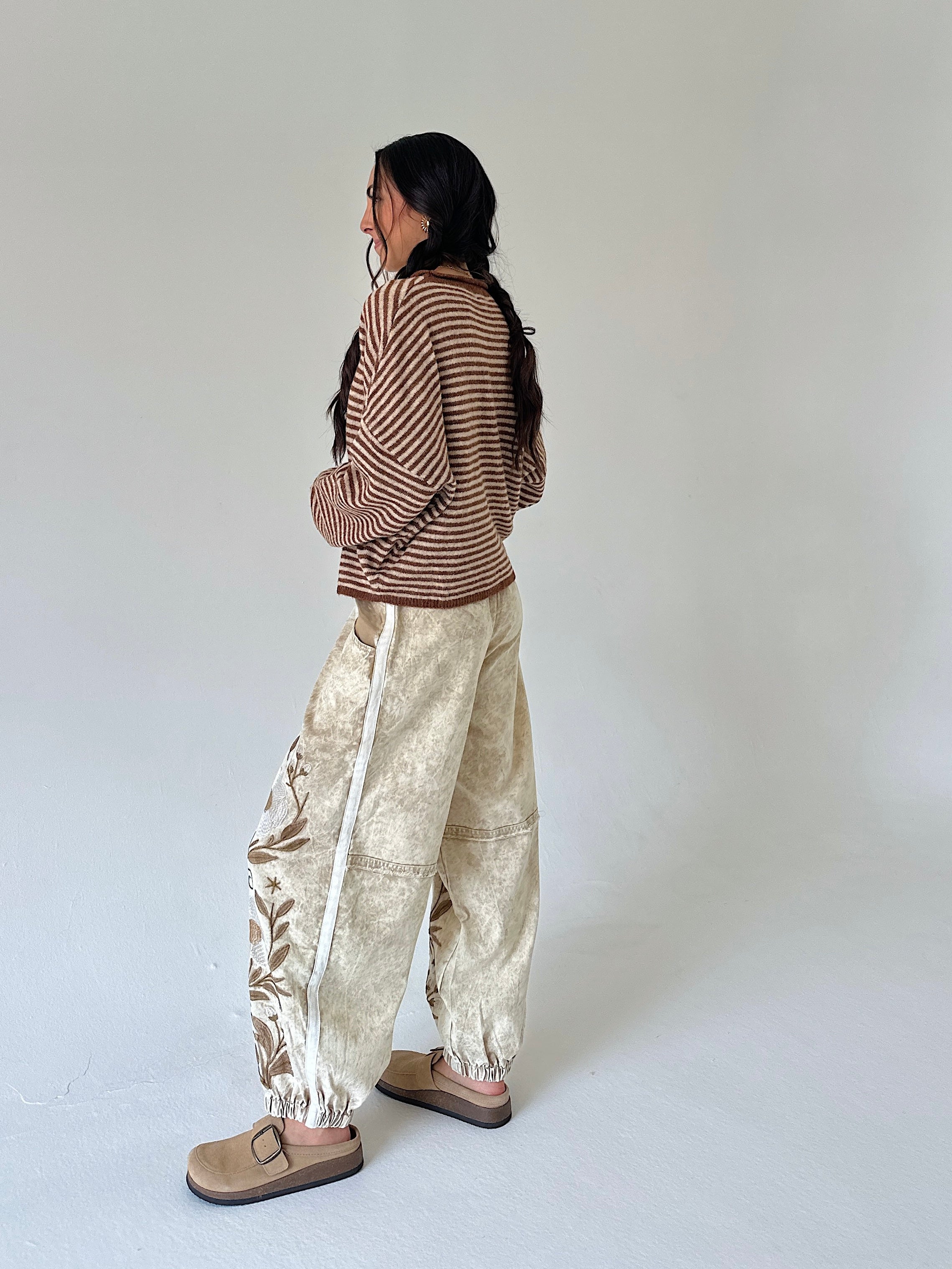 A woman stands in profile wearing a brown and beige striped long-sleeve top, beige patterned wide-leg pants with a white side stripe, and tan slip-on shoes against a plain light gray studio background.