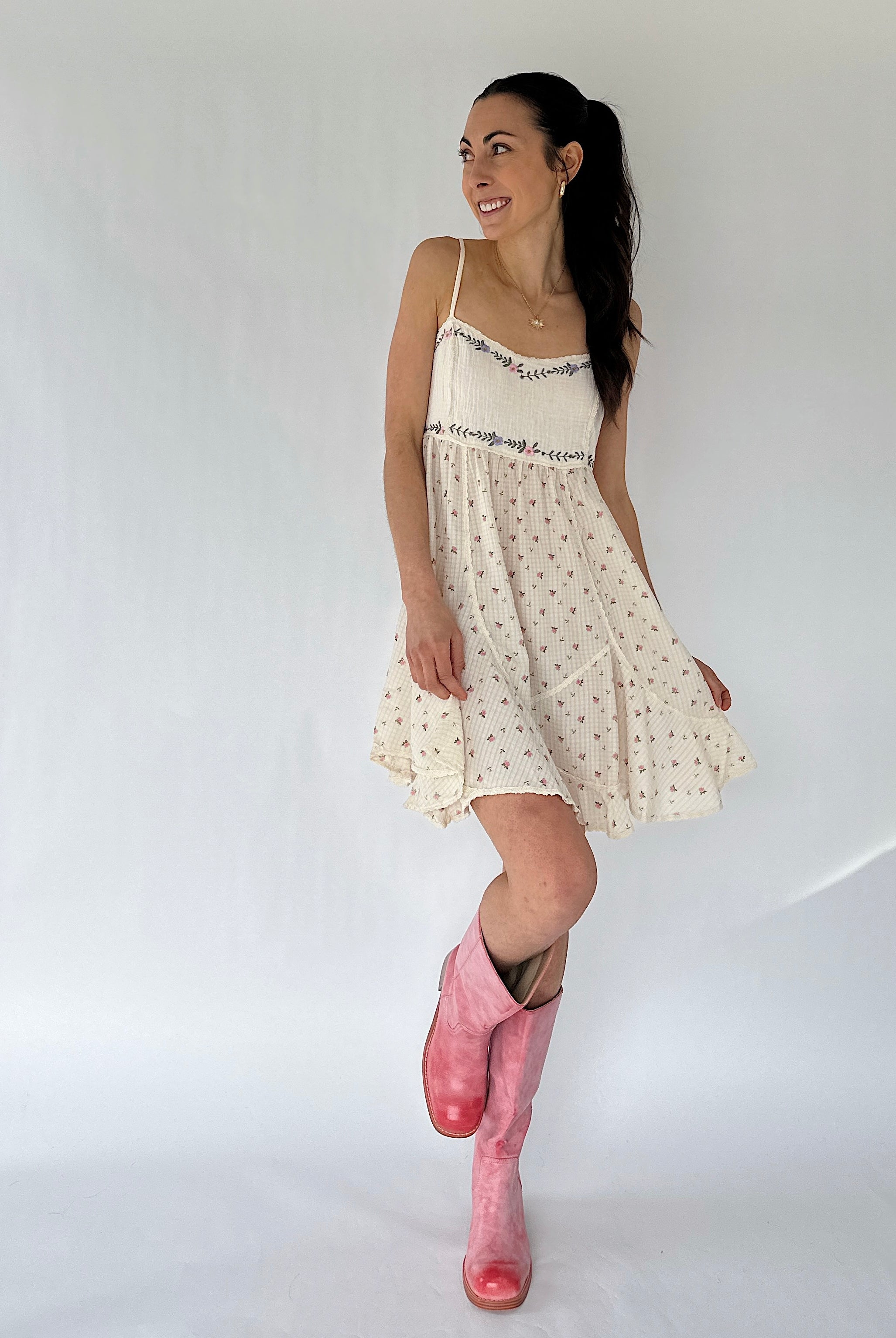 Woman wearing a white dress with floral patterns and pink boots on a plain background
