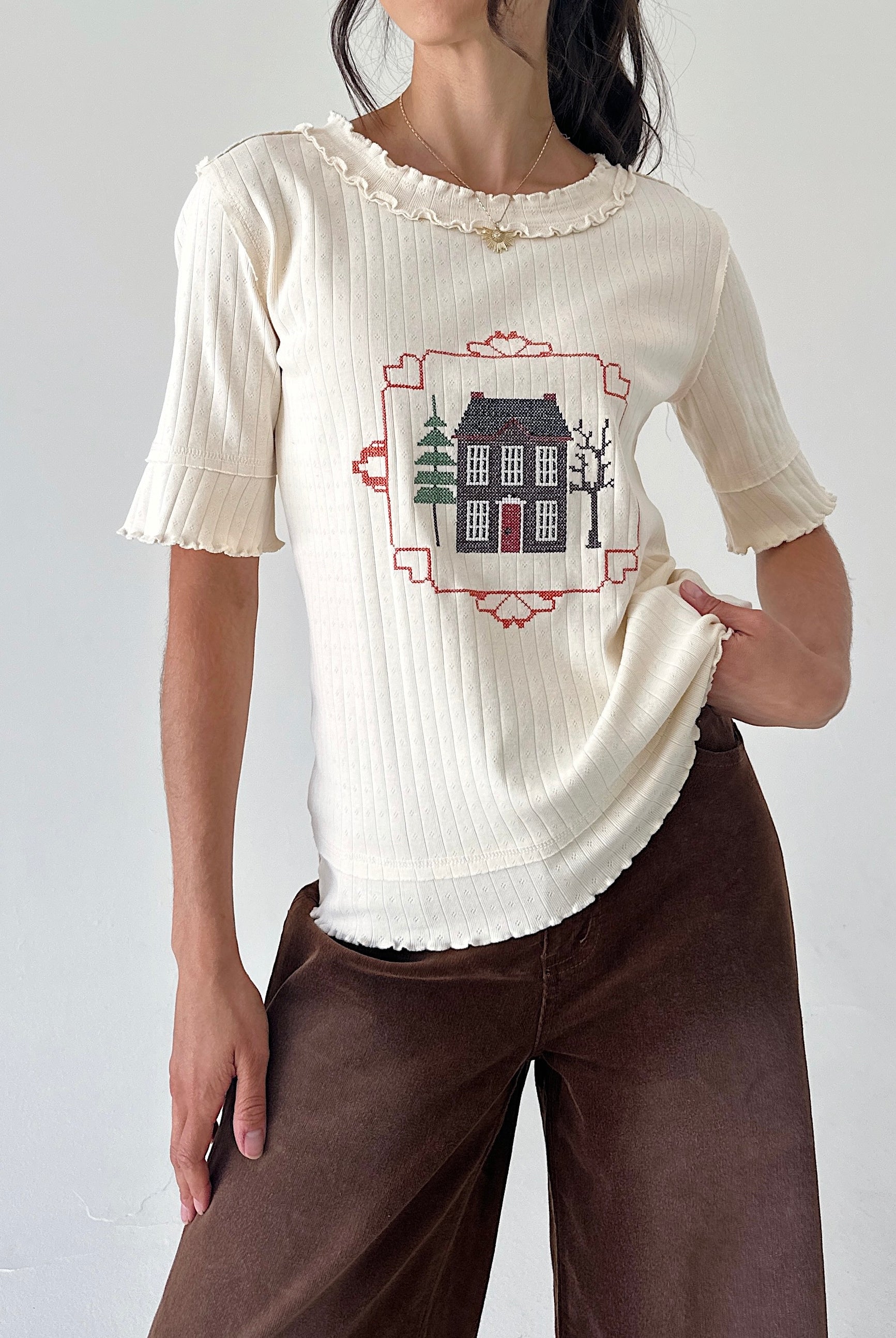 A woman wearing a cream ribbed short-sleeve top with a central house embroidery, paired with brown wide-leg pants, against a plain light gray background.
