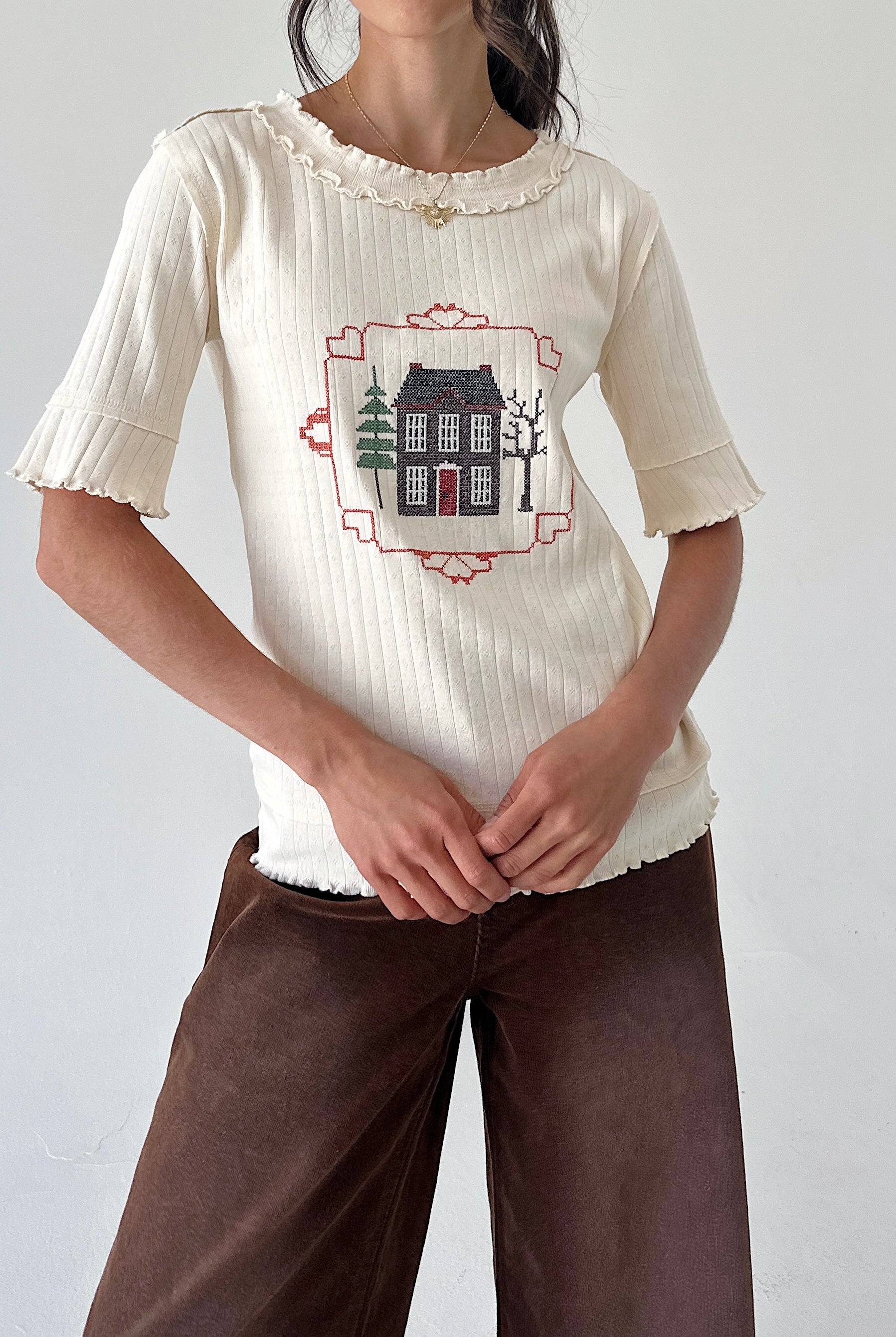 A woman wearing a cream ribbed short-sleeve top with a ruffled neckline and a central embroidered house graphic, paired with brown pants, against a plain light gray wall.