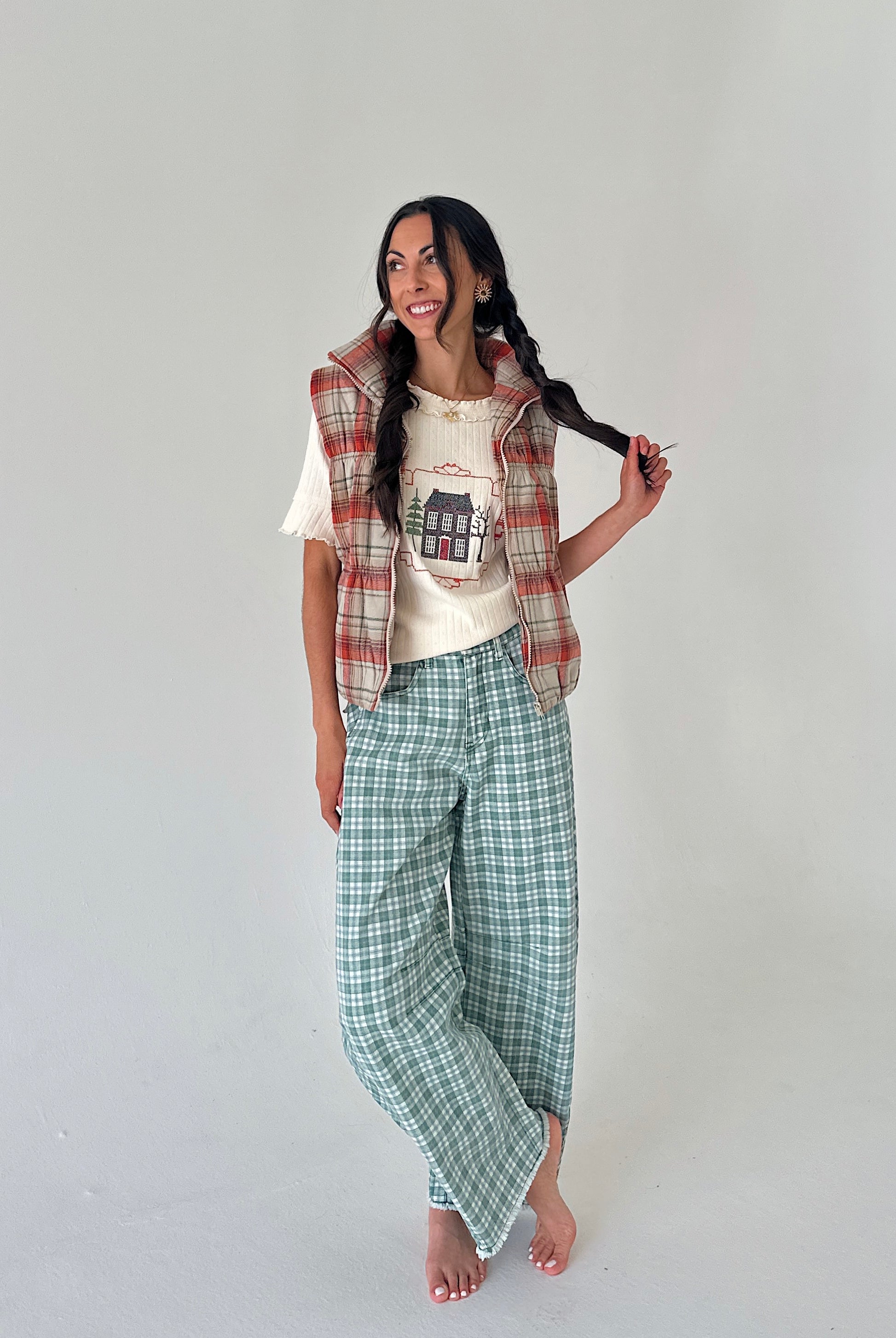 A woman stands barefoot against a plain light gray studio background, wearing a cream graphic T-shirt, a red and beige plaid sleeveless vest, and green-and-white plaid cropped pants.
