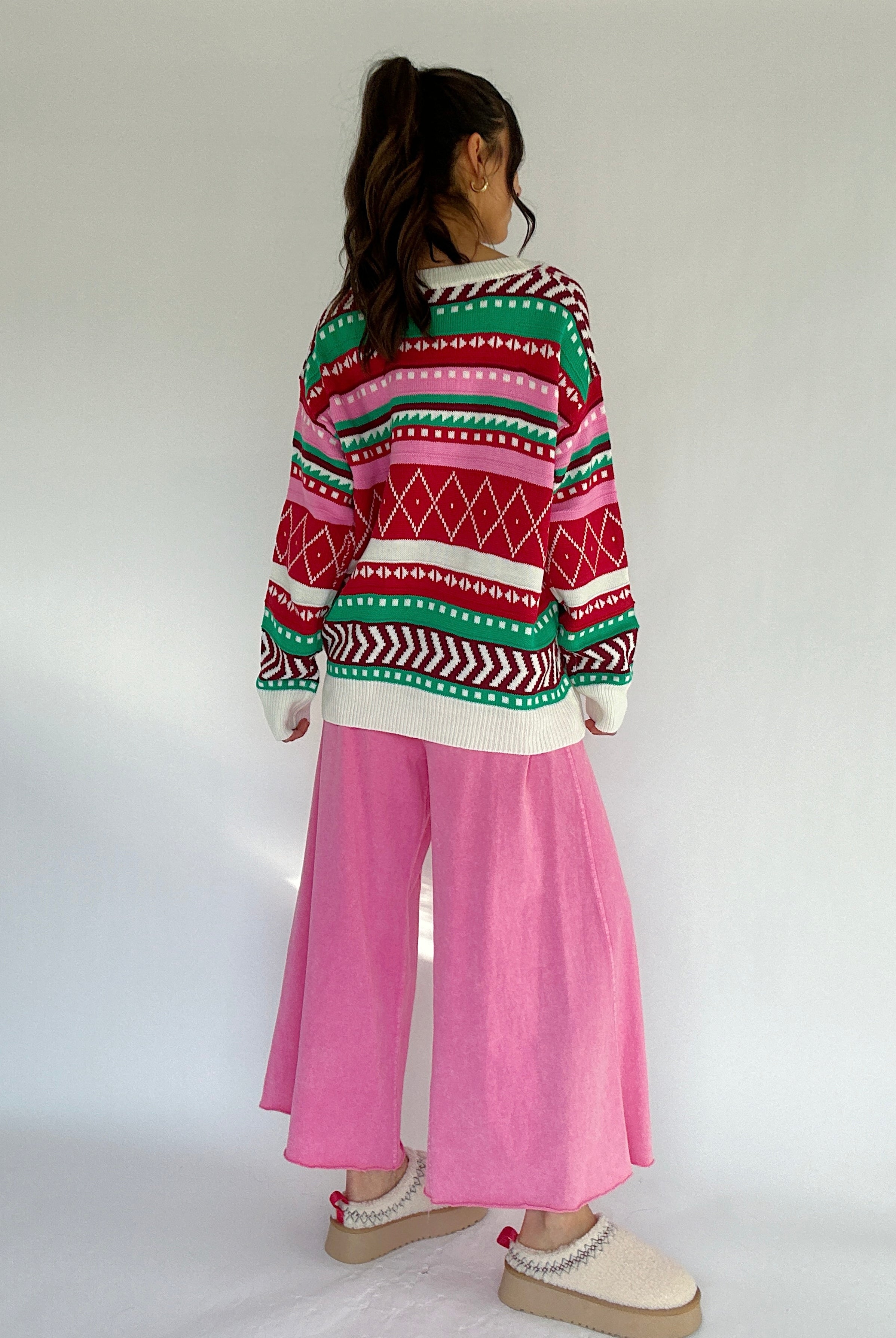 Person wearing a colorful patterned sweater and pink pants on a white background from back