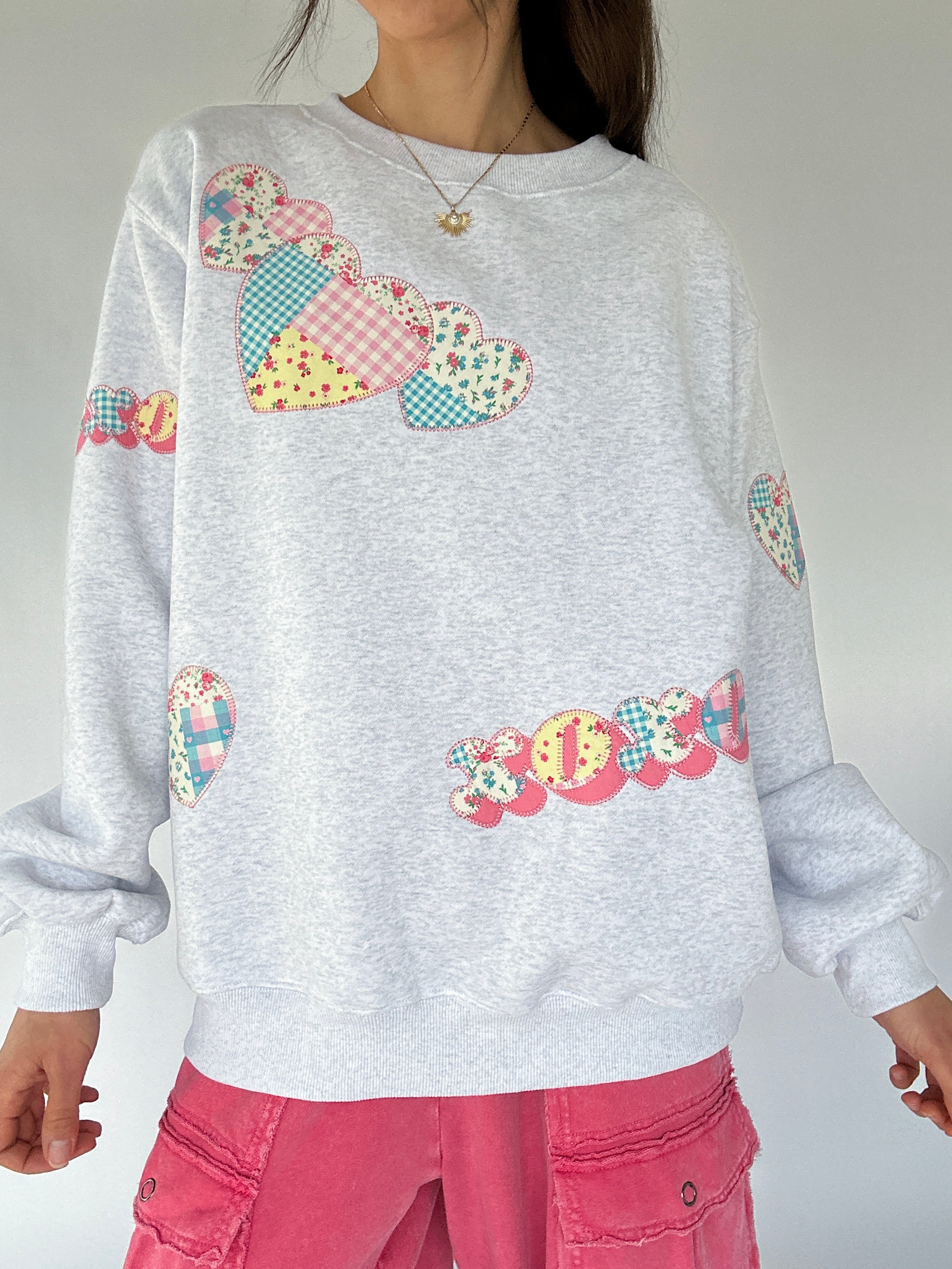 Person wearing a light gray sweatshirt with colorful patch xoxo and heart designs on a plain background