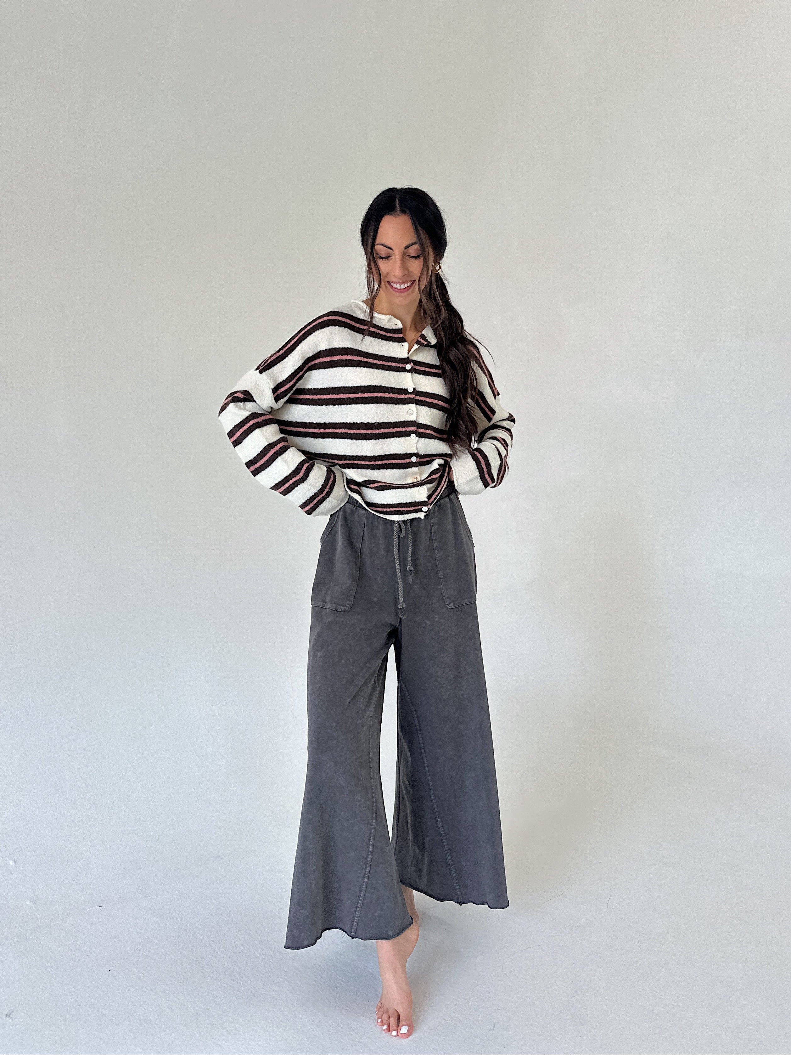 A woman stands barefoot in a studio setting wearing a cream knit top with dark brown horizontal stripes and dark gray wide-leg pants.