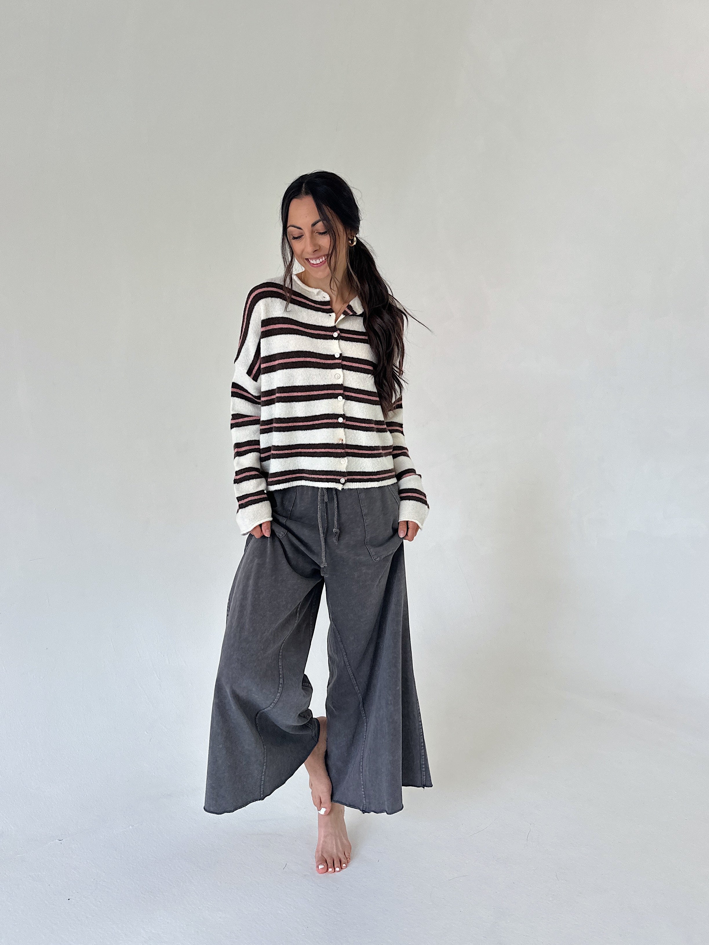 A woman stands barefoot in a minimalist white studio setting, wearing a cream and dark brown horizontally striped knit sweater and loose dark gray wide-leg pants.