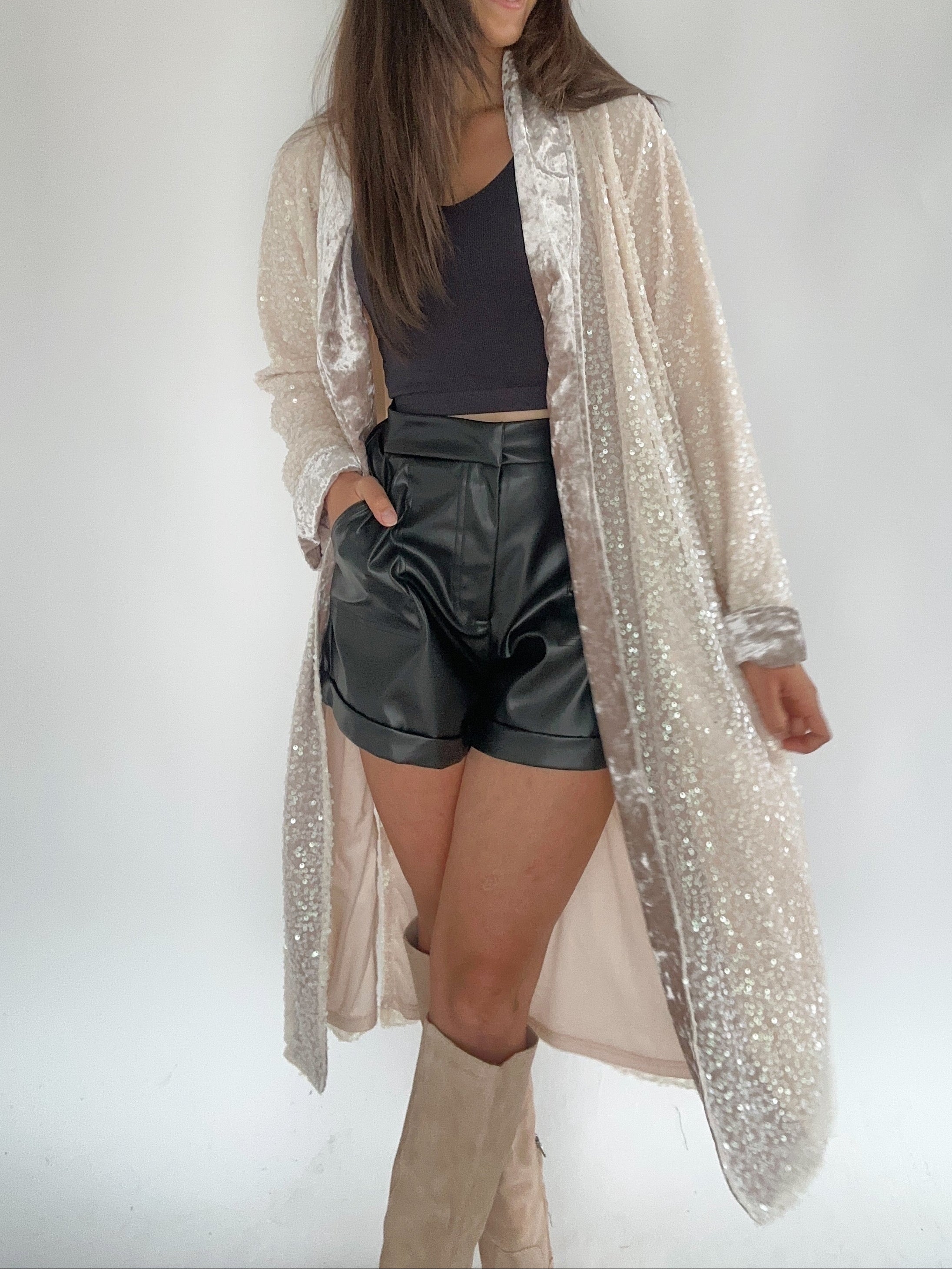 A woman wearing a beige velvet duster with sequin embellishments over a black top and denim shorts, accessorized with a wide-brimmed hat.