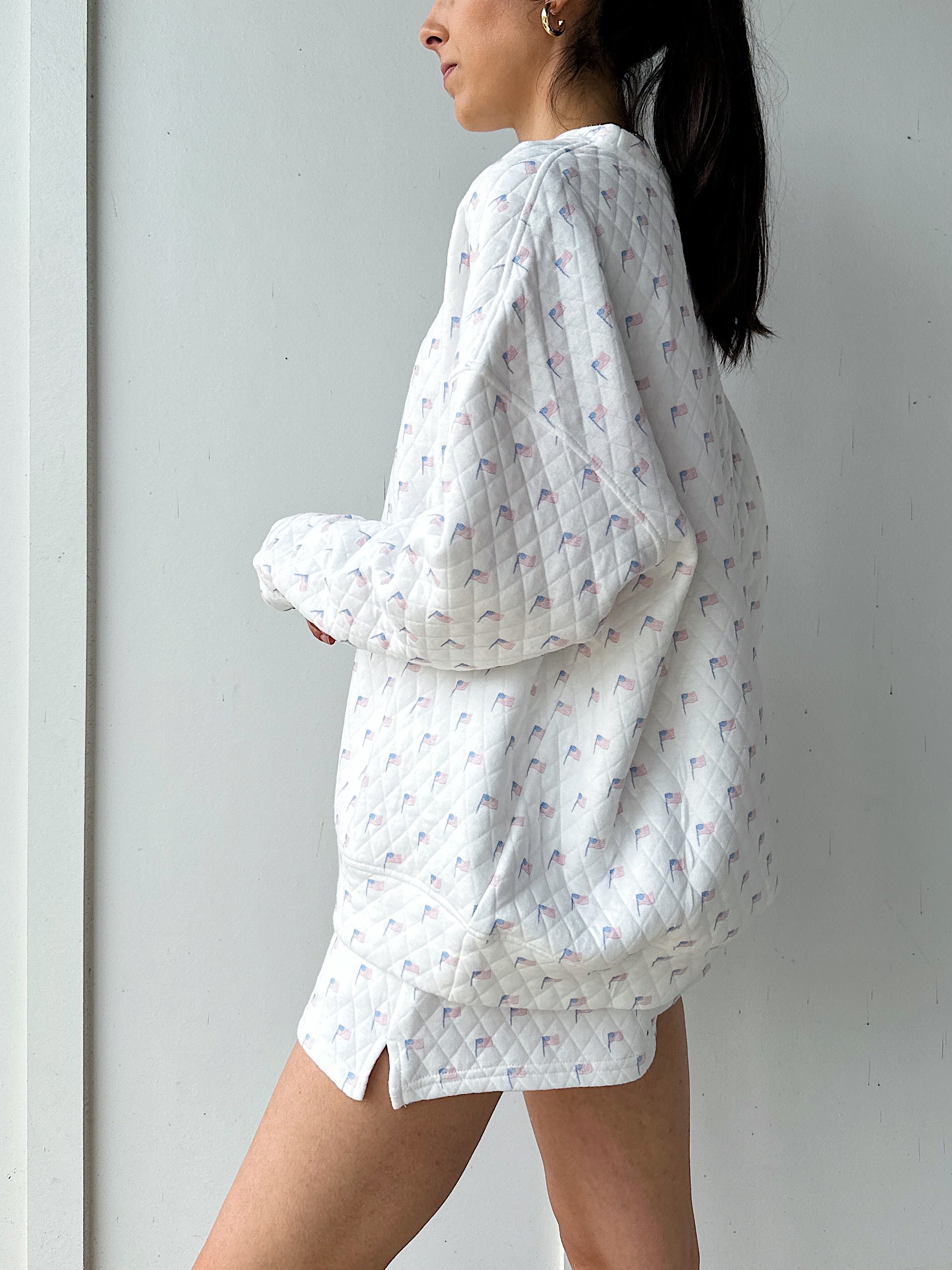 Woman wearing a white quilted flag patterned sweatshirt against a plain background