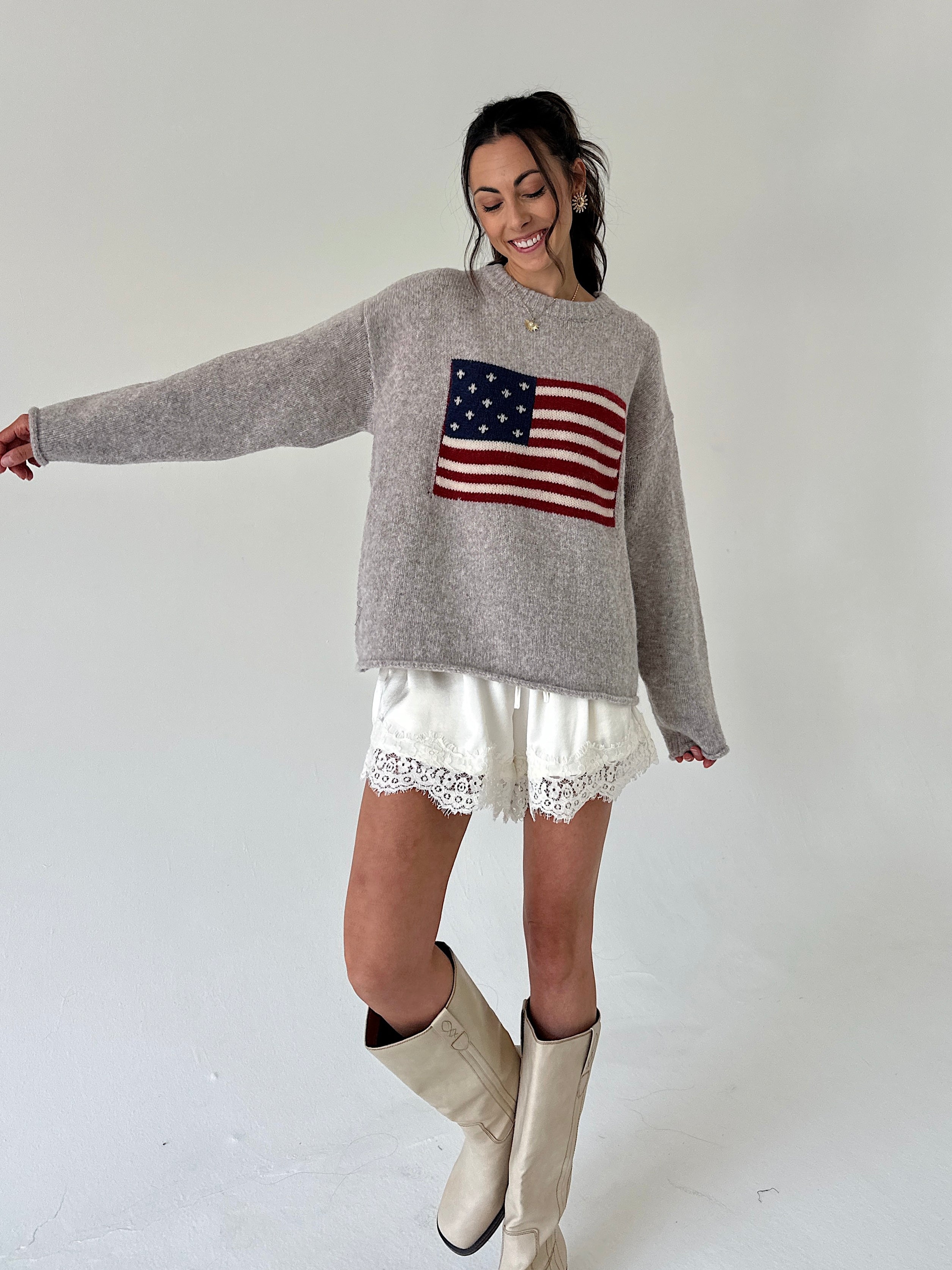 A woman modeling a light gray knit sweater with an American flag graphic on the chest, paired with white lace-trim shorts and beige cowboy boots, against a plain light gray studio background.