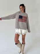 A woman modeling a light gray knit sweater with an American flag graphic on the chest, paired with white lace-trim shorts and beige cowboy boots, against a plain light gray studio background.