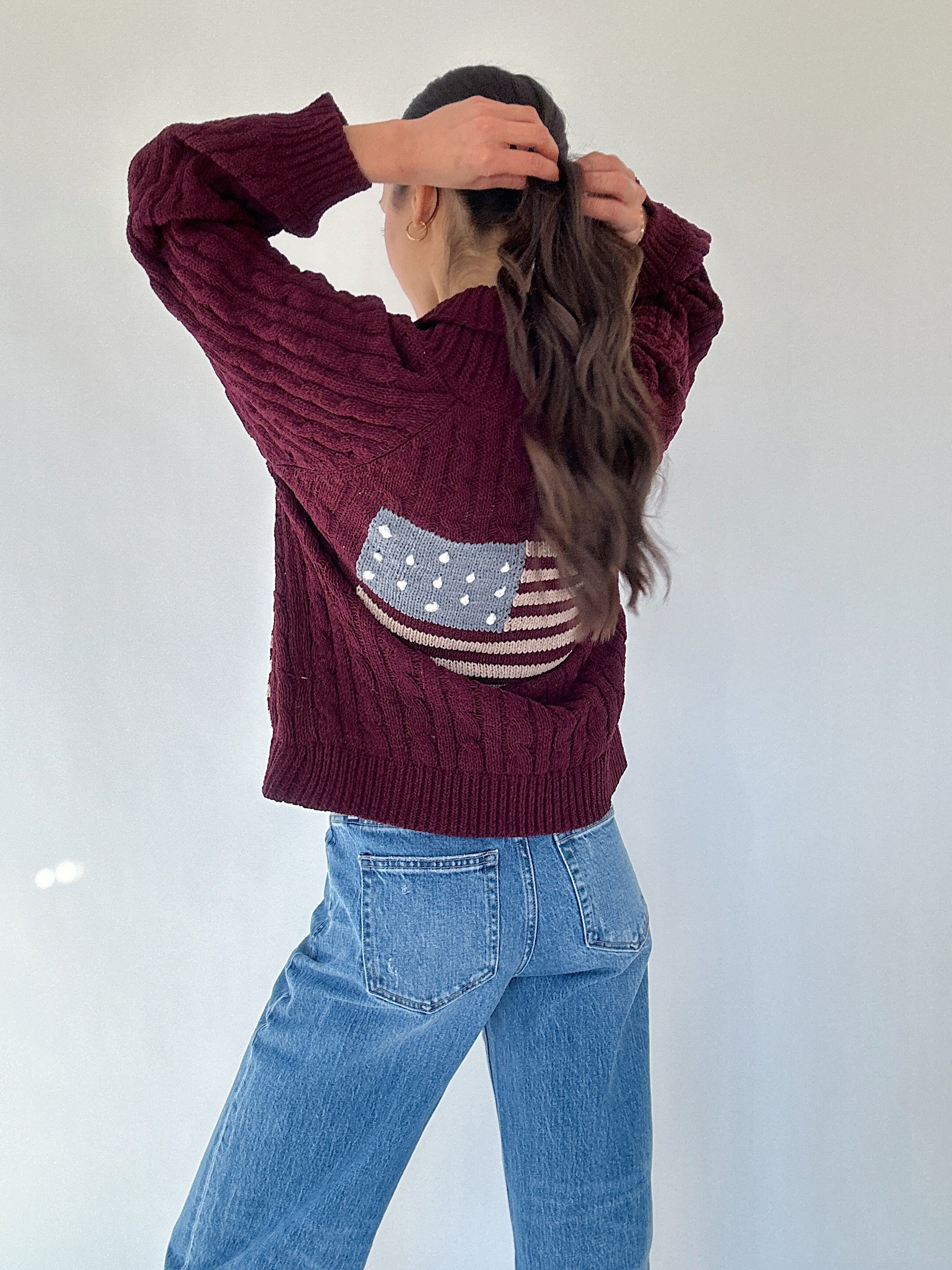 Person wearing a maroon sweater with an American flag design, standing against a white background holding hair