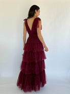 Woman wearing a burgundy evening gown with a sheer overlay on a white background showing back