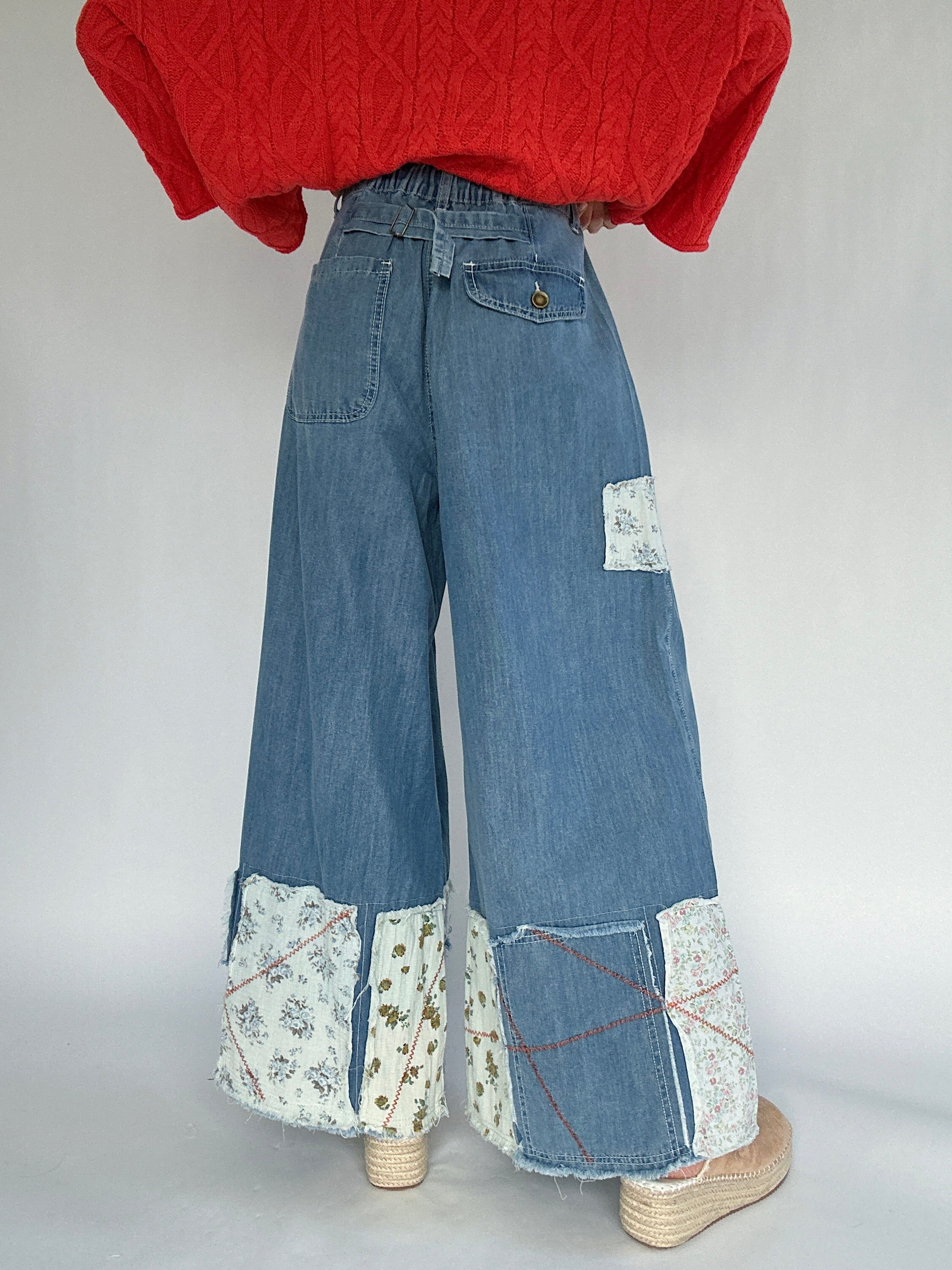 Person wearing a coral sweater and blue jeans with patchwork details a back buckle and back pockets on a plain background