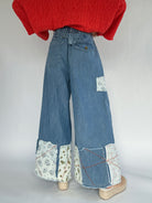 Person wearing a coral sweater and blue jeans with patchwork details a back buckle and back pockets on a plain background