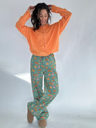 Woman wearing an orange cardigan sweater and green fall patterned pants on a white background