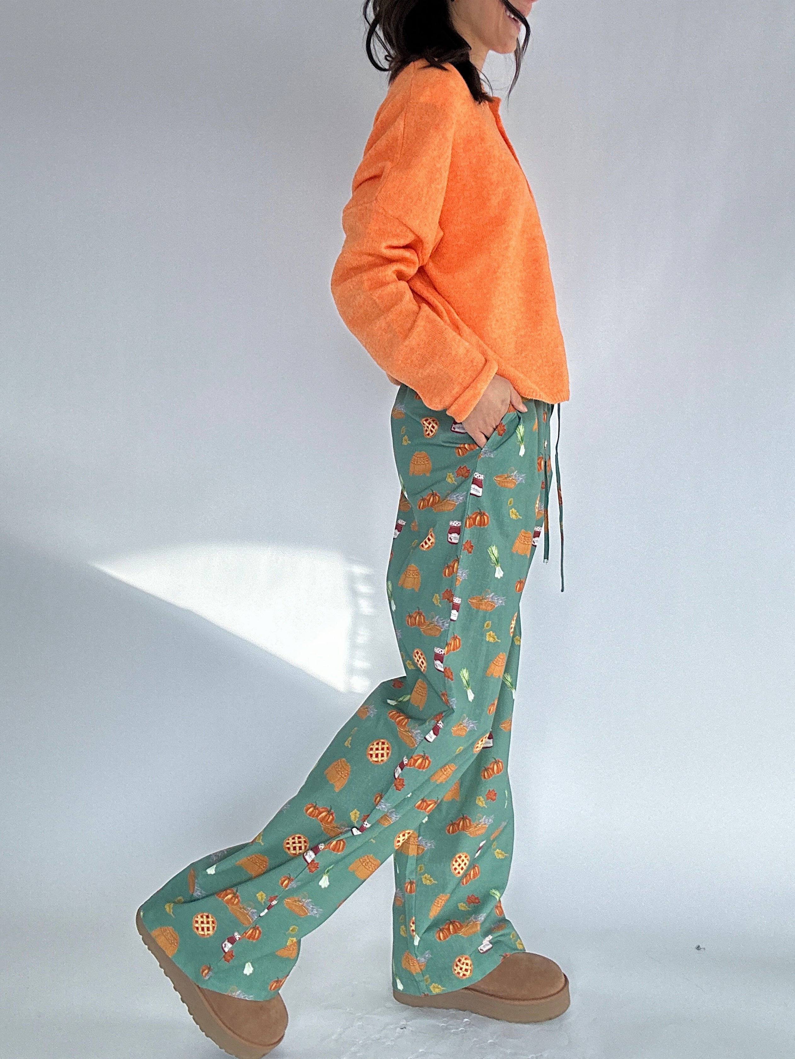Person wearing an orange cardigan top and green fall patterned pants on a white background showing side view with pockets