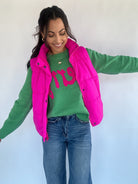 Woman wearing green sweater with text under a hot pink puffer vest and jeans on a plain background