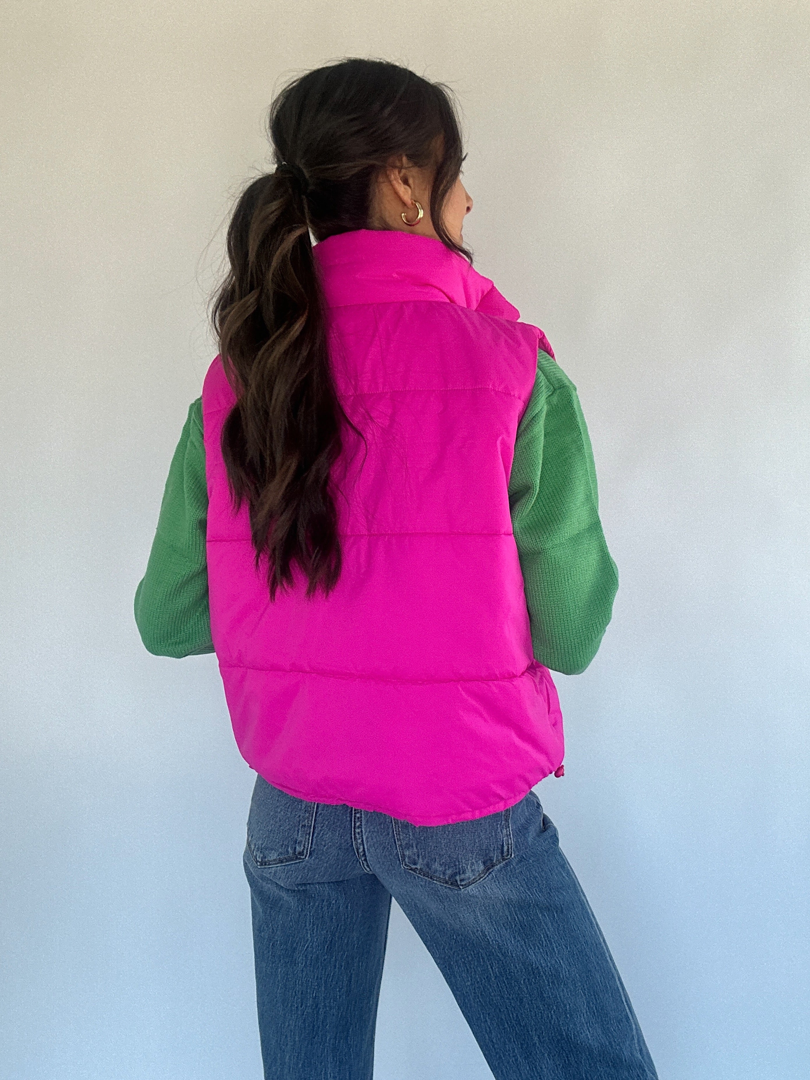 Hot pink puffer vest over a green sweater with jeans from the back on a plain background