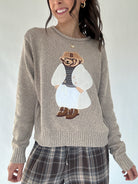 Person wearing a taupe sweater with a dressed up teddy bear embroidered character design on a plain background