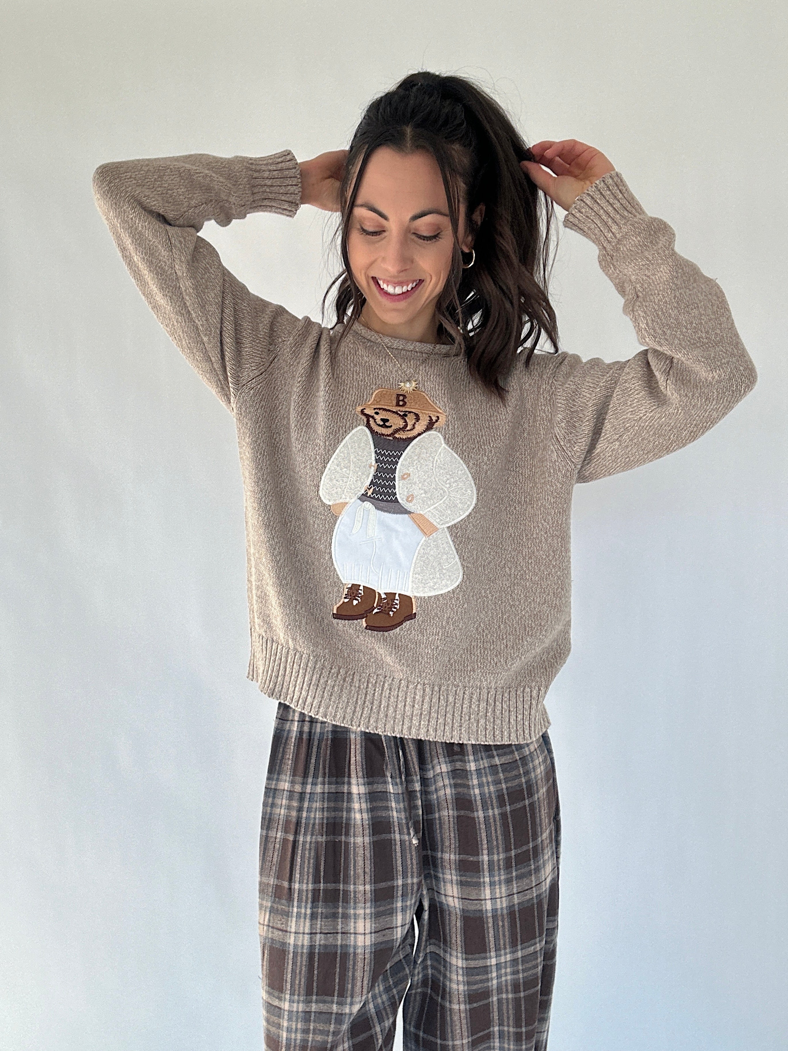 Woman wearing a beige sweater with a dressed up teddy bear character design on a plain background