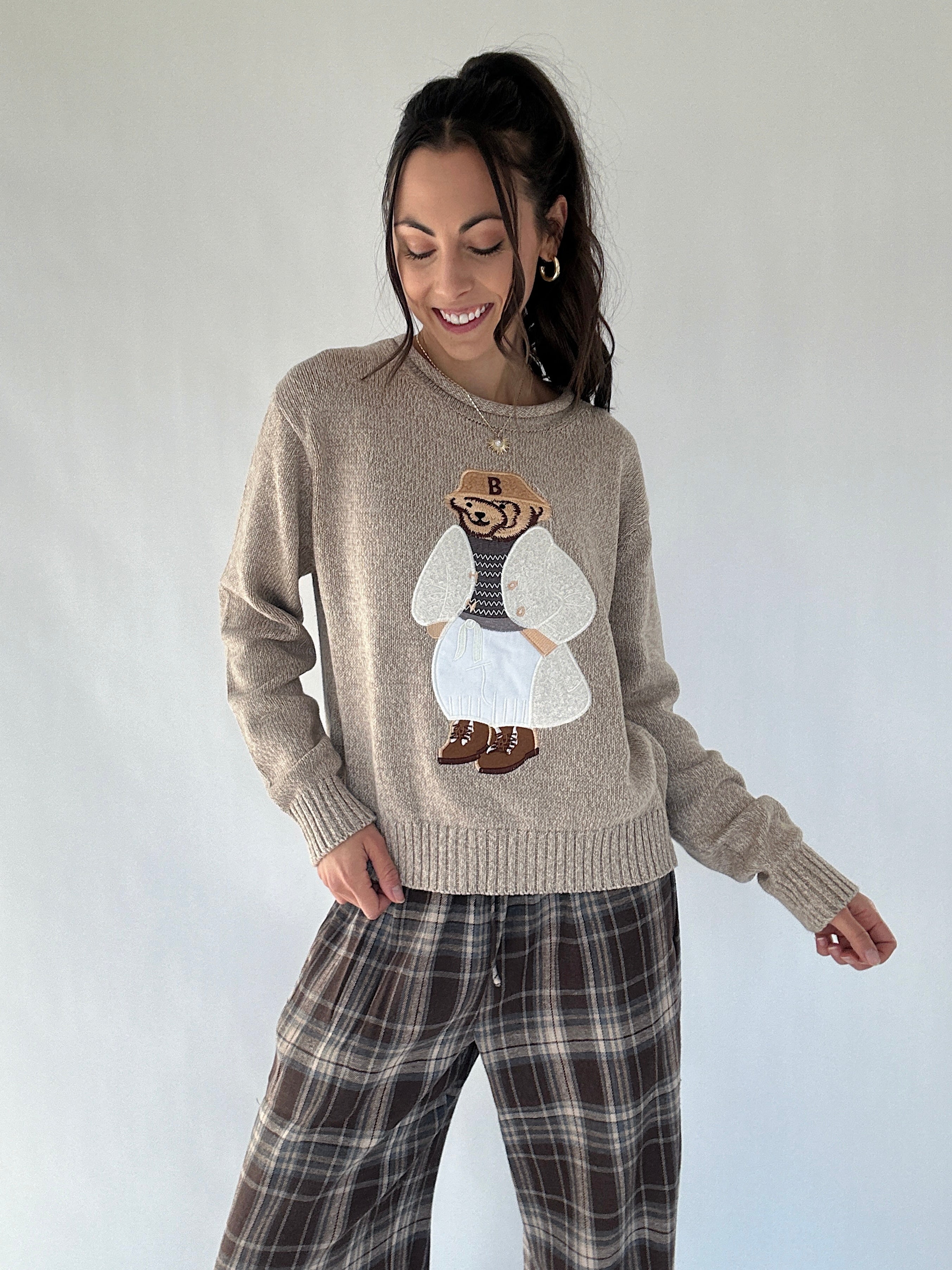 Woman wearing a beige sweater with a graphic design of a bear dressed up on a plain background