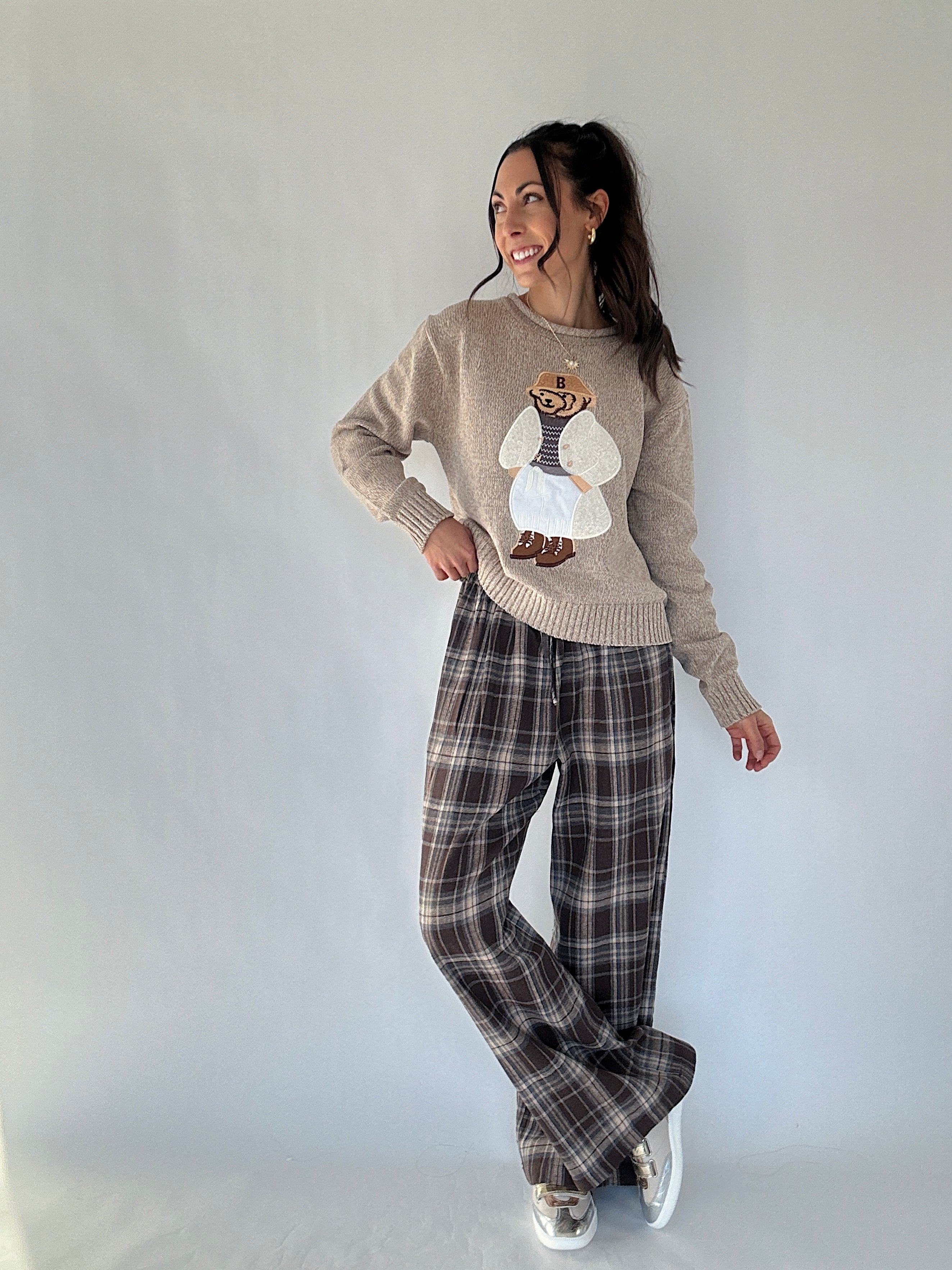 Woman wearing a sweater with a dressed up teddy bear embroidered design and plaid pants against a white background