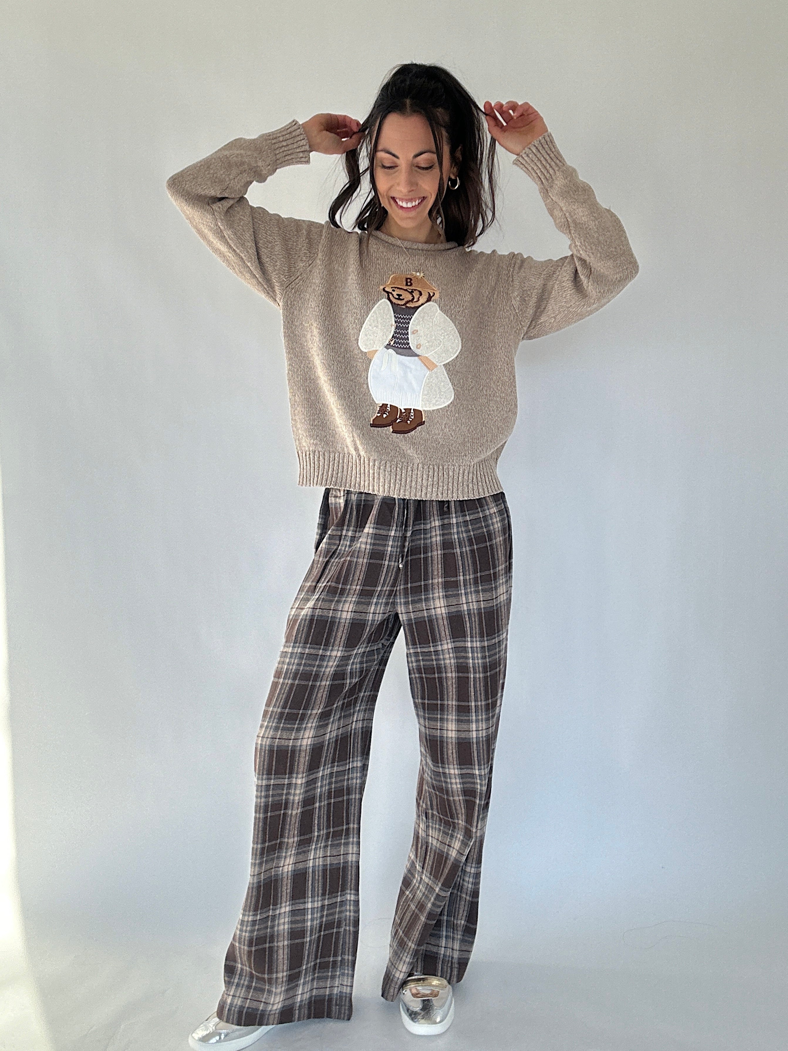 Person wearing a beige sweater with a teddy bear design and plaid pants on a white background
