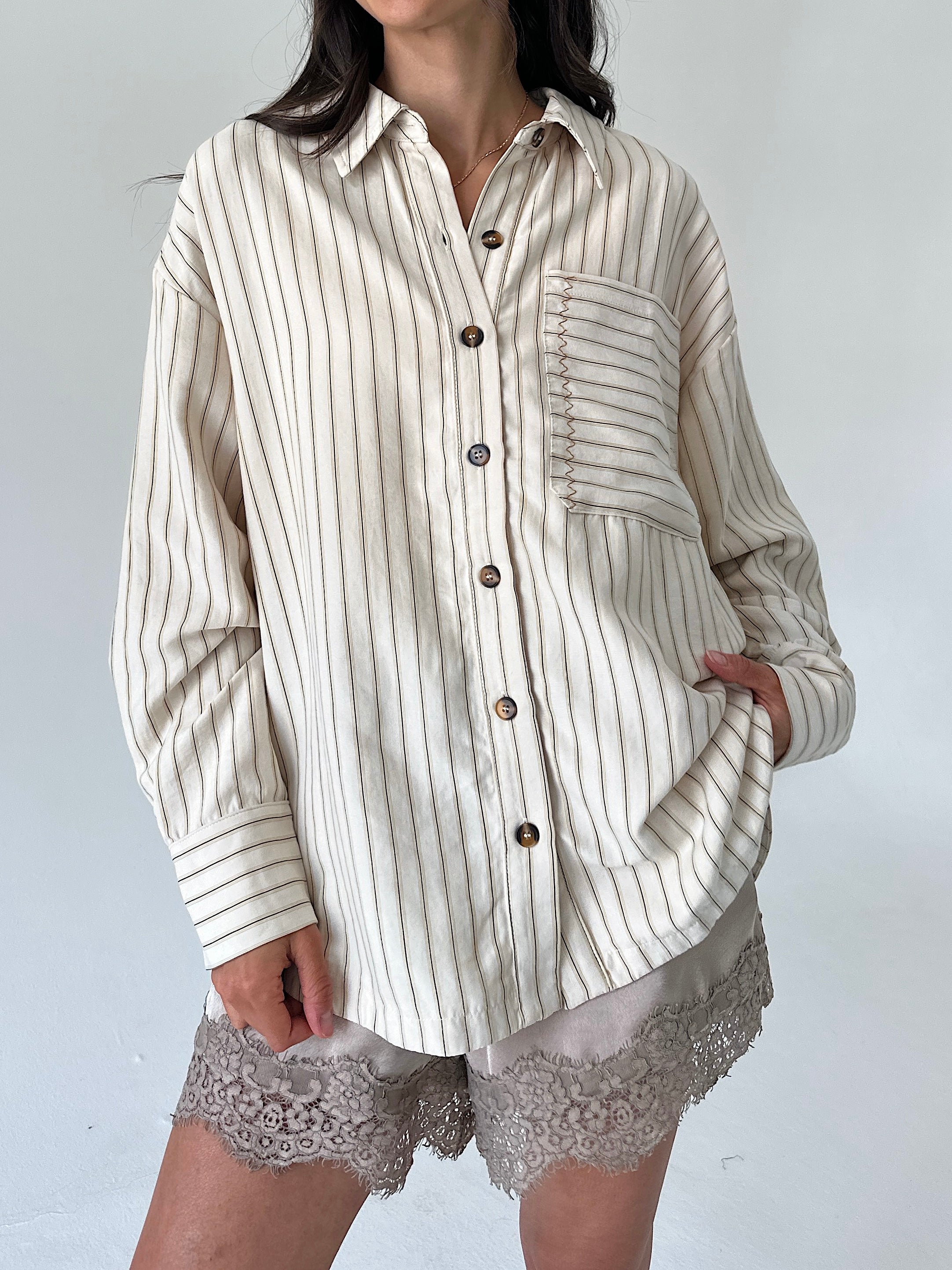 A woman wearing an oversized cream-colored button-up shirt with thin vertical brown stripes, paired with lace-trimmed shorts, against a plain light gray background.