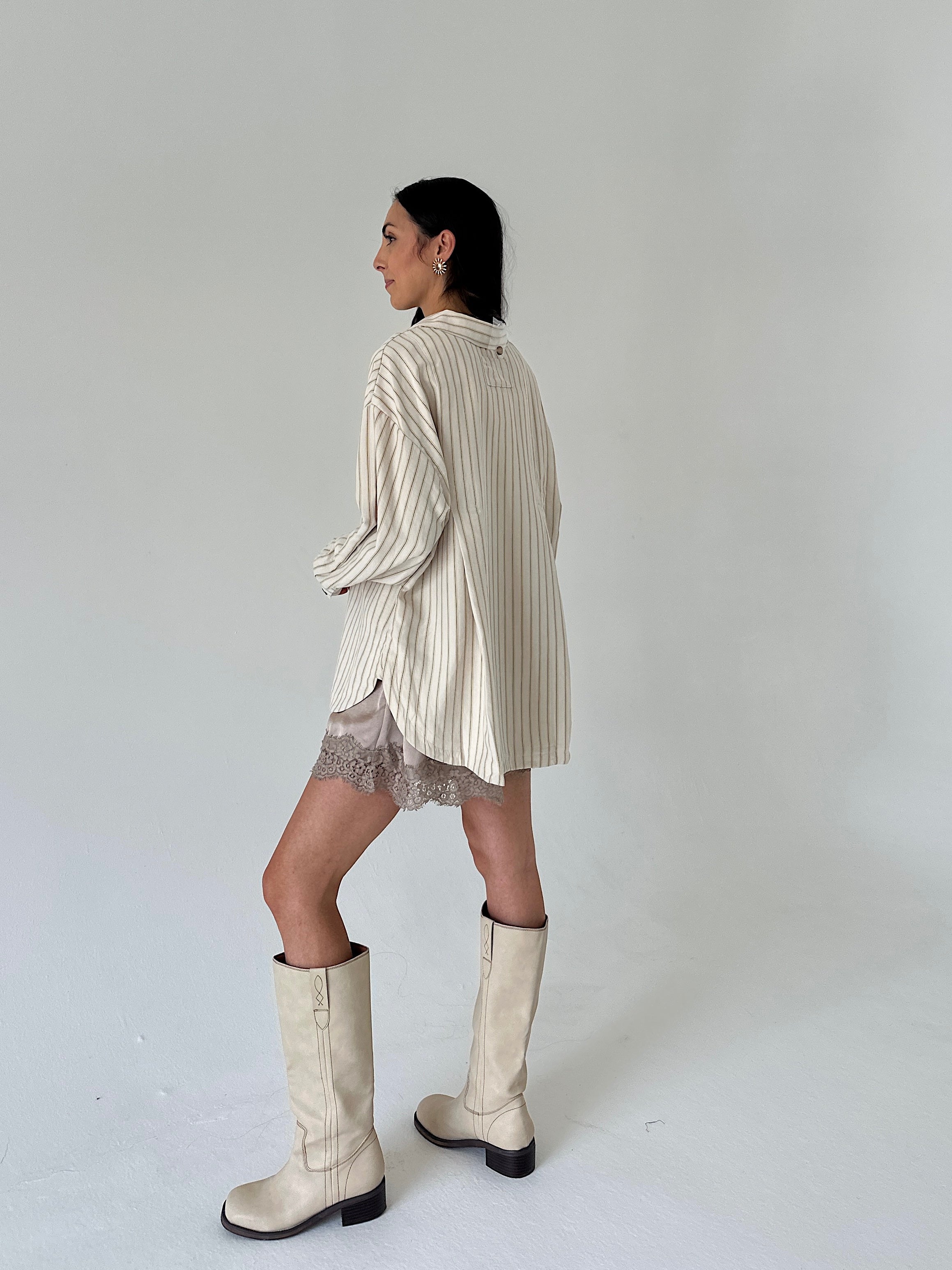 A woman is shown from the back wearing a cream and beige vertically striped oversized shirt dress, a sheer lace mini skirt, and off-white knee-high boots against a plain light gray studio background.
