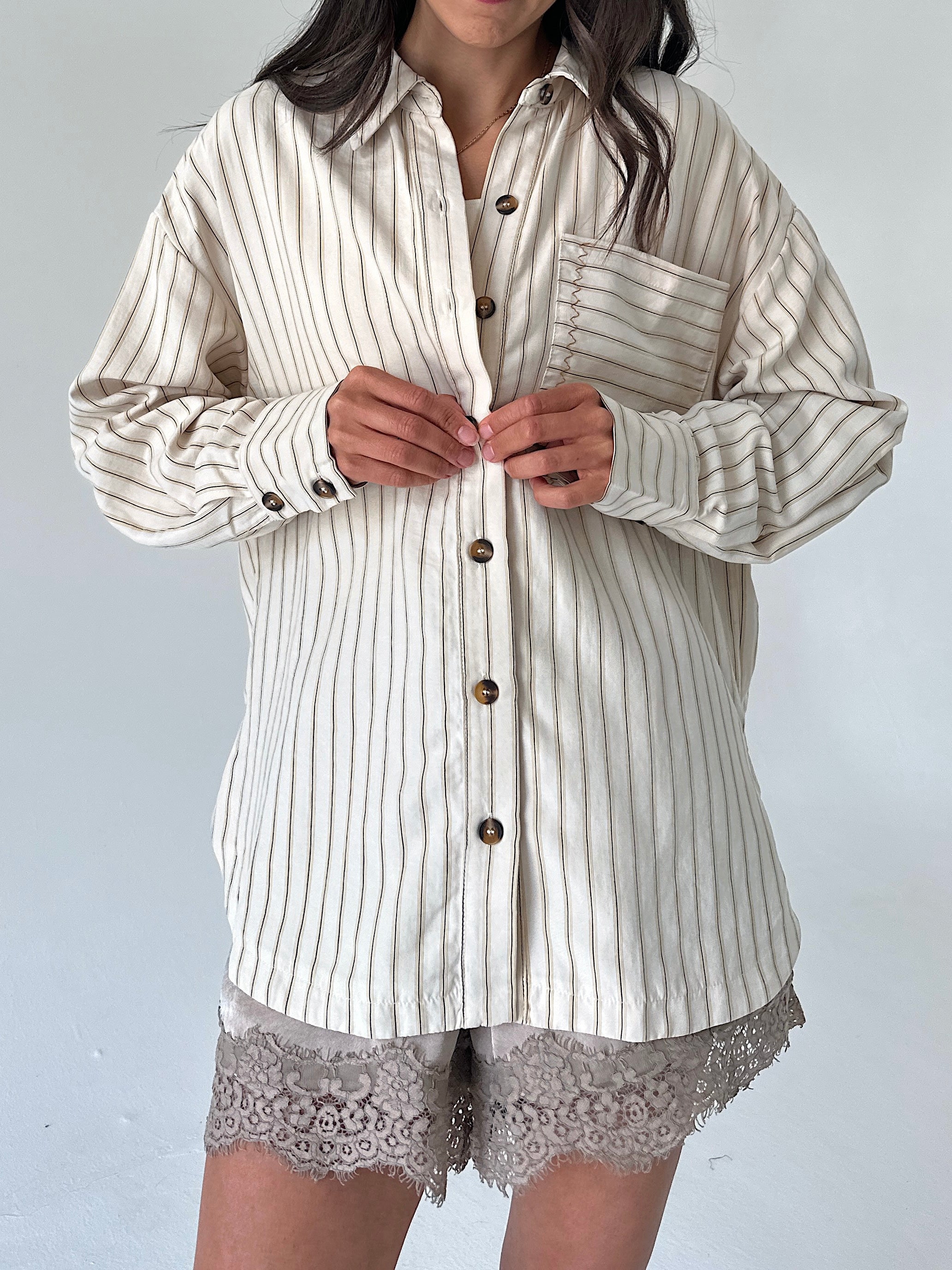 A woman wearing an oversized cream-colored button-up shirt with thin vertical brown stripes, paired with lace-trimmed shorts, against a plain light gray background.