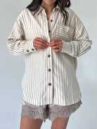 A woman wearing an oversized cream-colored button-up shirt with thin vertical brown stripes, paired with lace-trimmed shorts, against a plain light gray background.