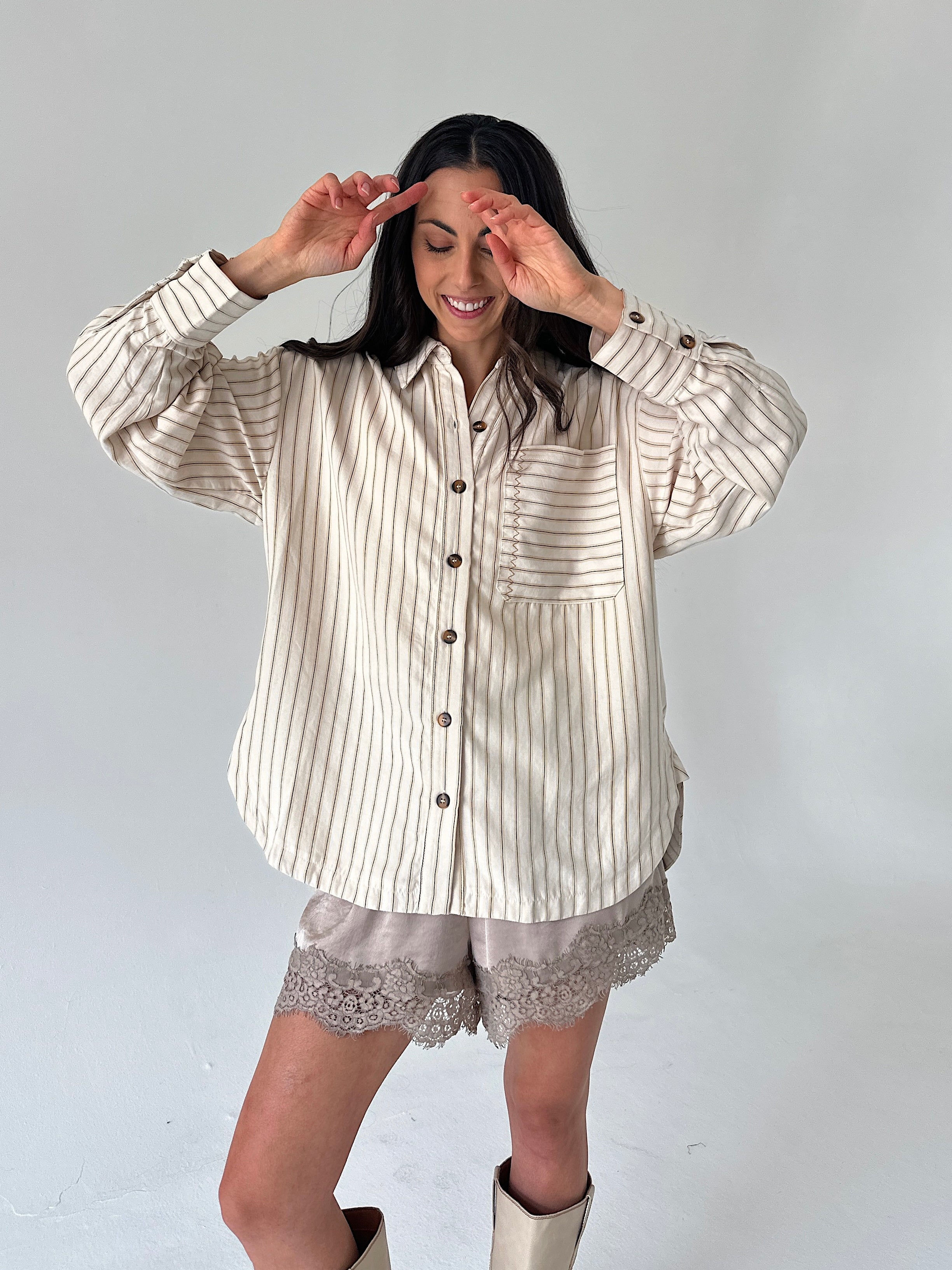 A woman modeling a cream and brown vertically striped button-up shirt with long sleeves, paired with matching lace-trim shorts and beige boots, against a plain light gray studio background.