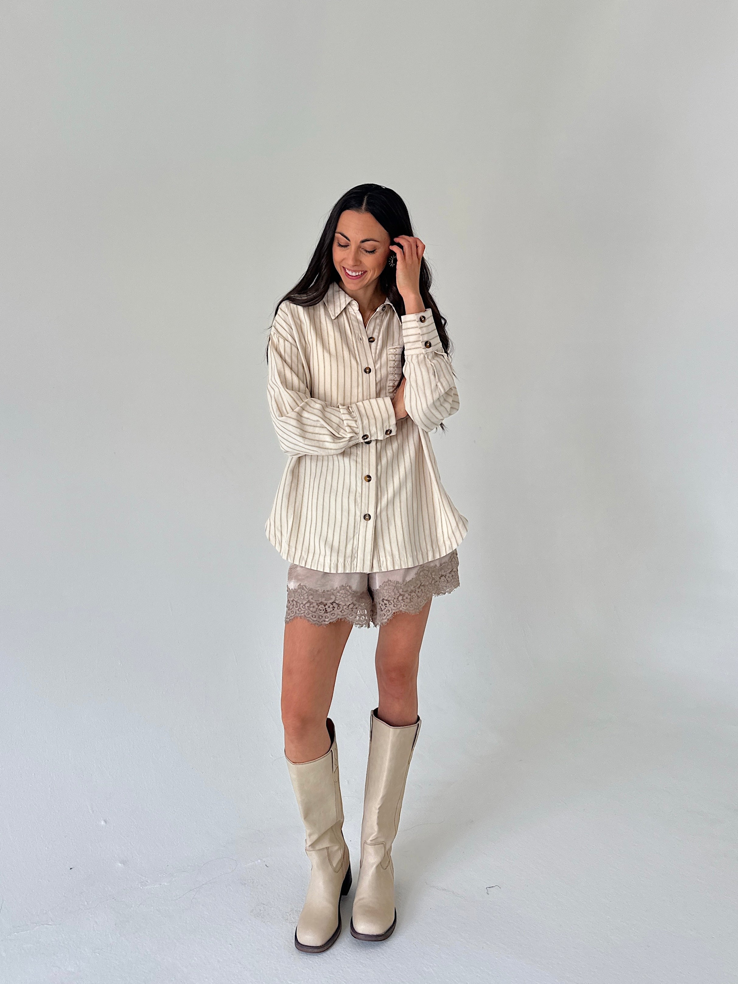 A woman modeling a cream and beige vertically striped button-down shirt with lace shorts and beige knee-high boots, standing against a plain light gray studio background.