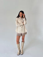 A woman modeling a cream and beige vertically striped button-down shirt with lace shorts and beige knee-high boots, standing against a plain light gray studio background.