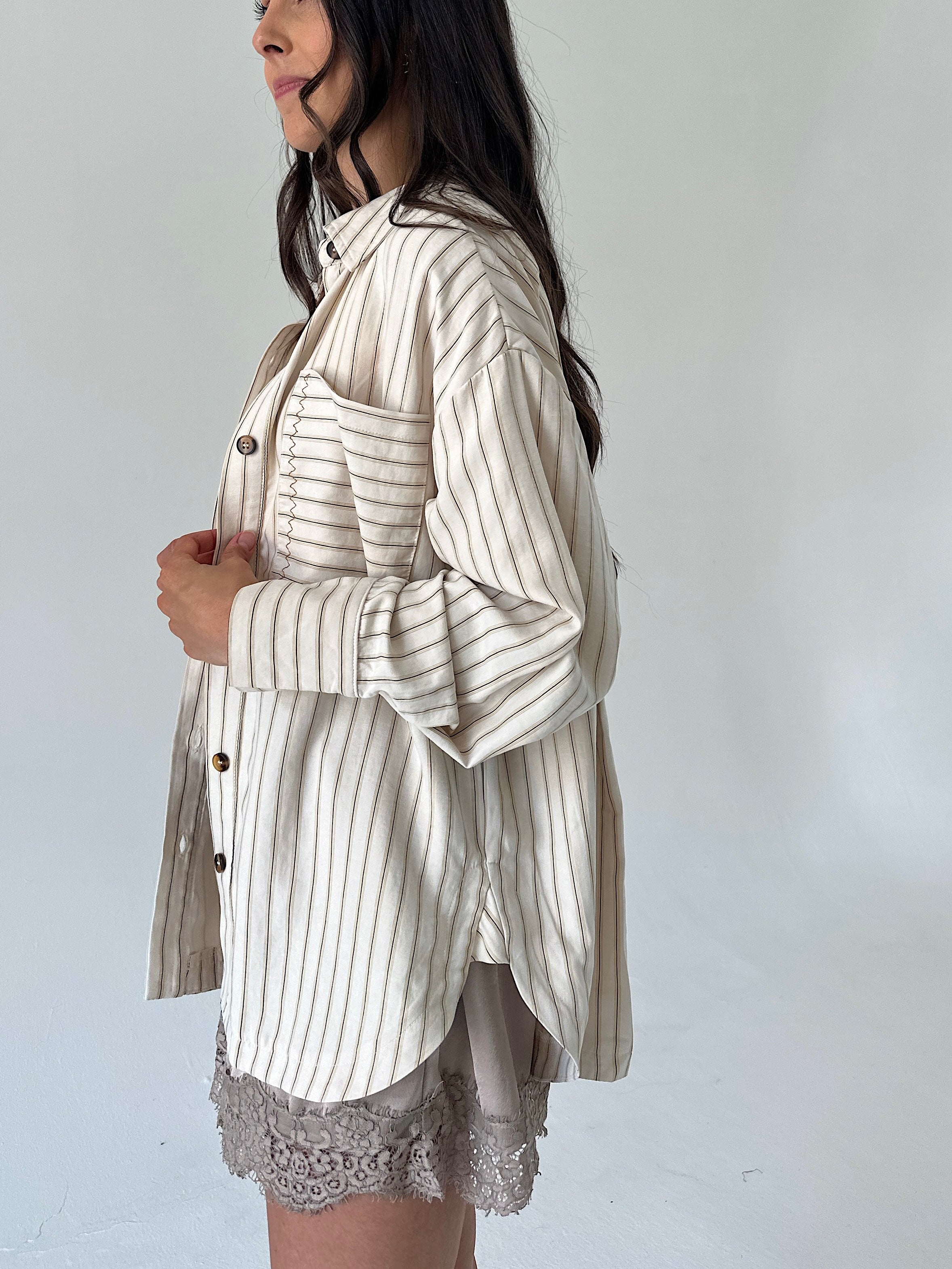 A woman wearing a cream-colored button-up shirt with thin vertical brown stripes, styled over a light gray lace-trimmed skirt, shown from a side profile against a plain light gray background.