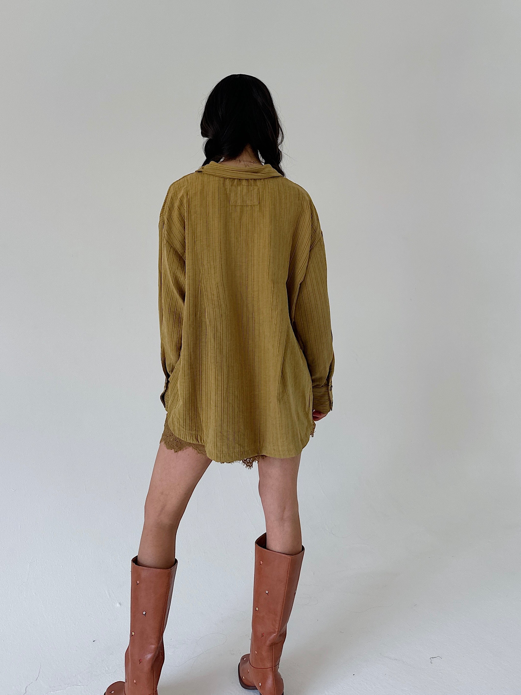 A model viewed from behind wearing a loose, olive-yellow shirt and brown leather knee-high boots against a plain white studio background.