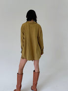A model viewed from behind wearing a loose, olive-yellow shirt and brown leather knee-high boots against a plain white studio background.
