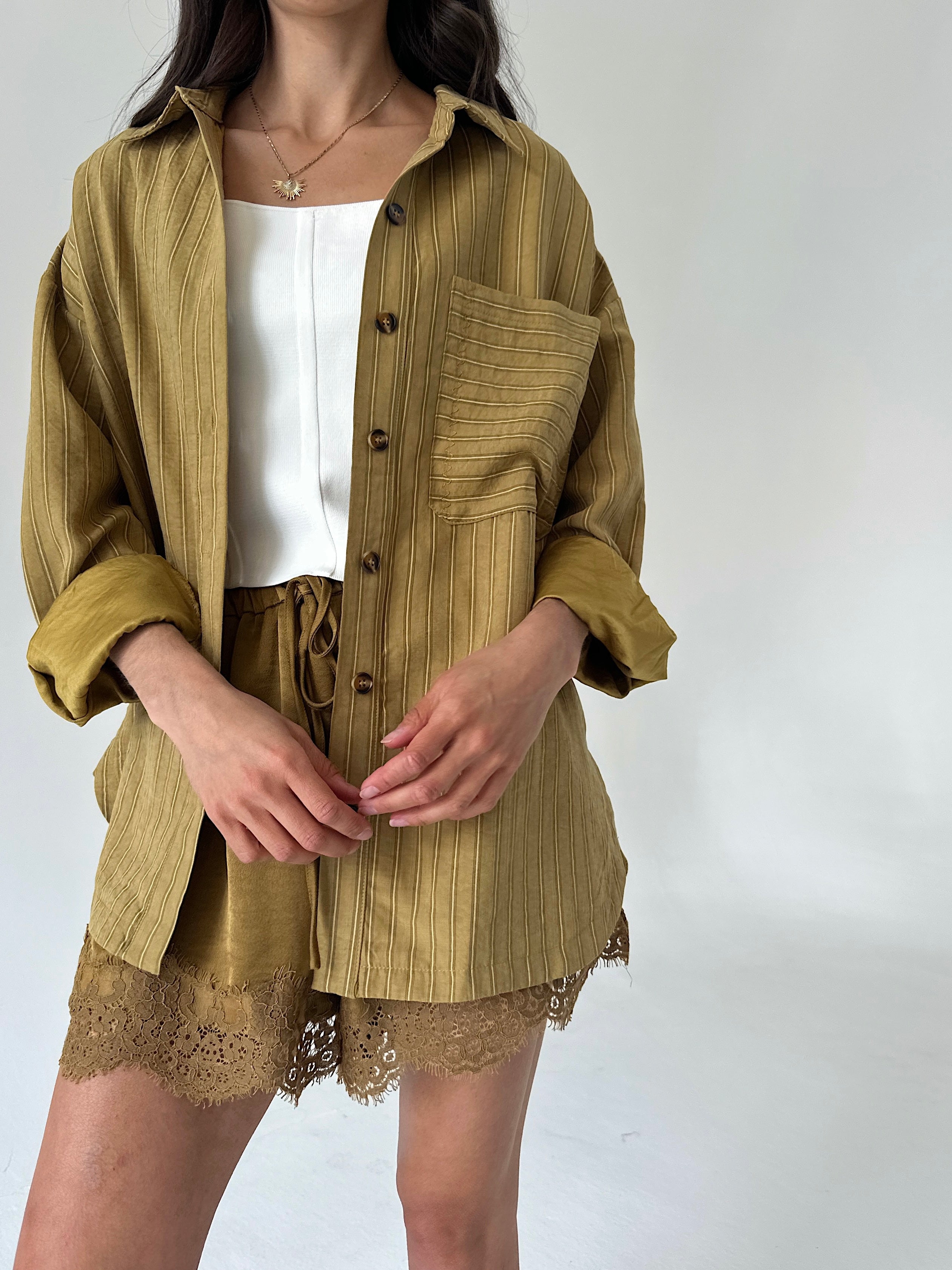 A model wearing a matching olive mustard button up shirt and shorts set with a white top underneath, photographed against a plain light gray background.
