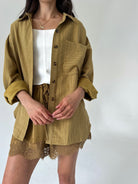 A model wearing a matching olive mustard button up shirt and shorts set with a white top underneath, photographed against a plain light gray background.