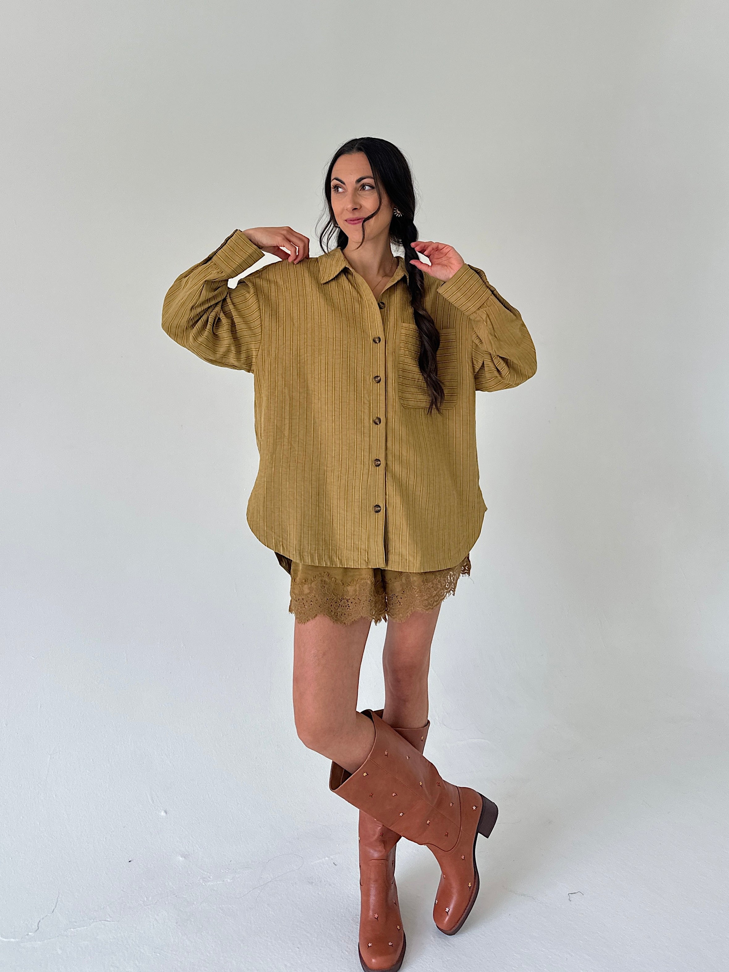 A woman modeling an oversized olive-brown linen button-up shirt with matching lace shorts and brown leather knee-high boots, standing against a plain light gray studio background.