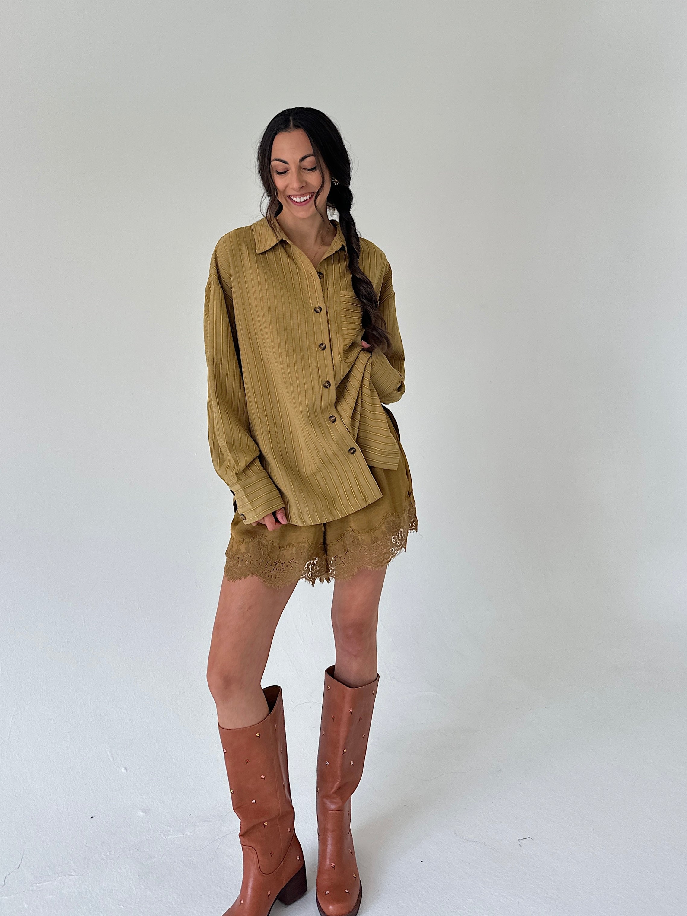 A woman modeling a matching olive mustard button-up shirt and lace shorts set, paired with brown leather knee-high boots, against a plain light gray studio background.