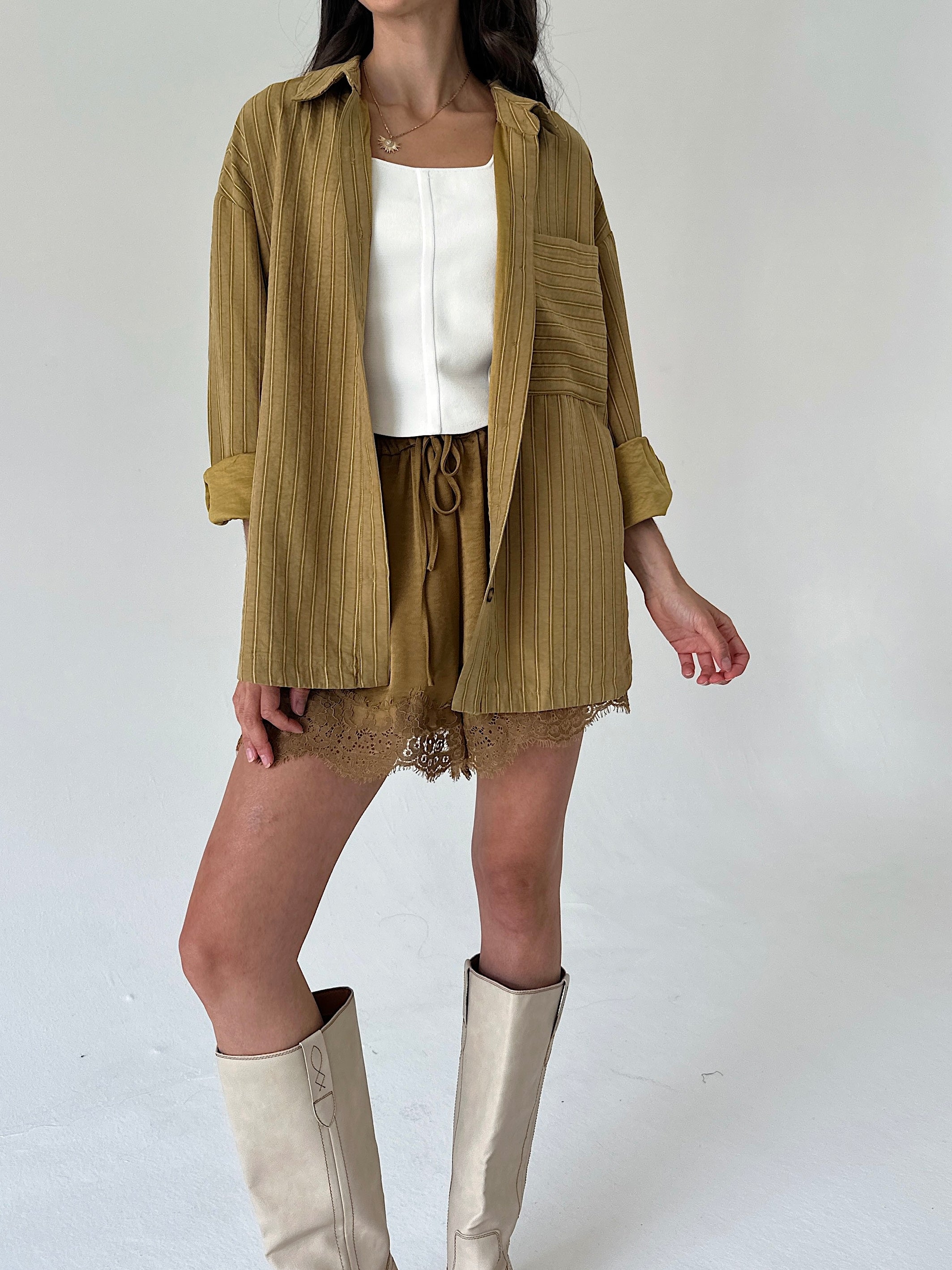 A model wearing a mustard yellow shirt jacket over a white top, paired with matching satin shorts and beige knee-high boots, against a plain light gray background.