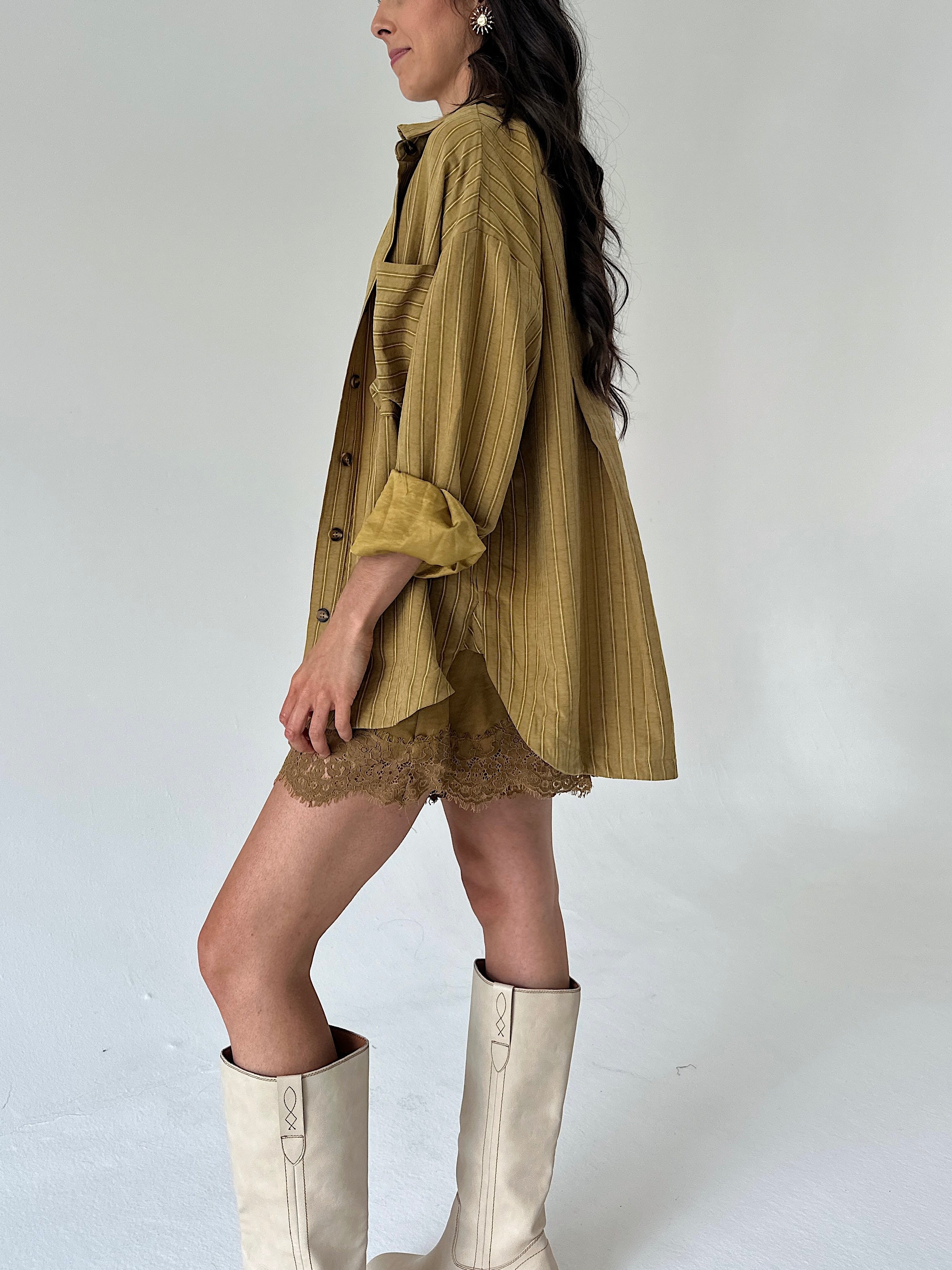 A woman wearing an oversized mustard yellow shirt with a lace hem shorts, styled with cream-colored knee-high boots against a plain light gray studio background.